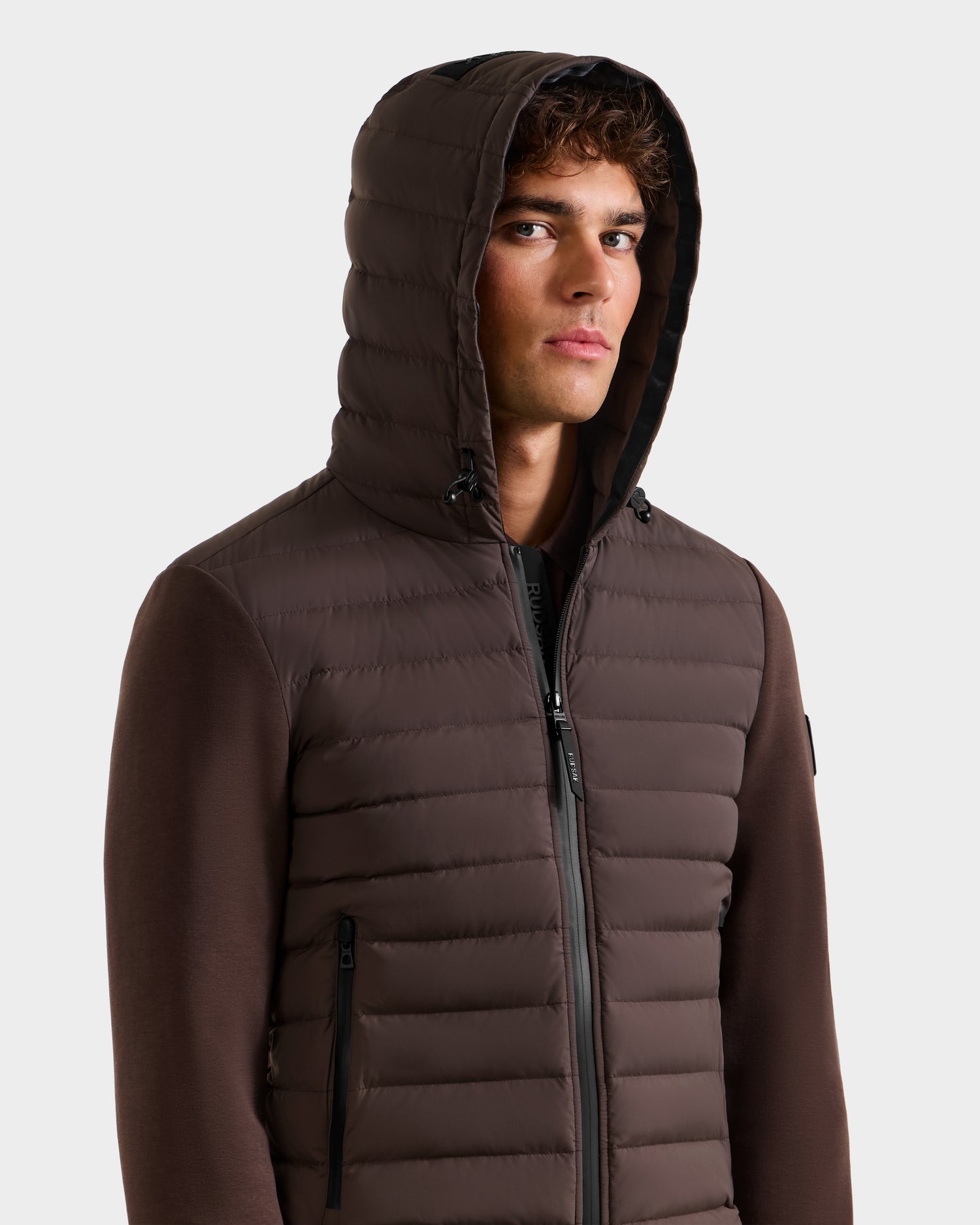 A young man faces forward against a white background, wearing the Rudsak DEE MAT - 6124028 Espresso men's lightweight hybrid down jacket with black accents.
