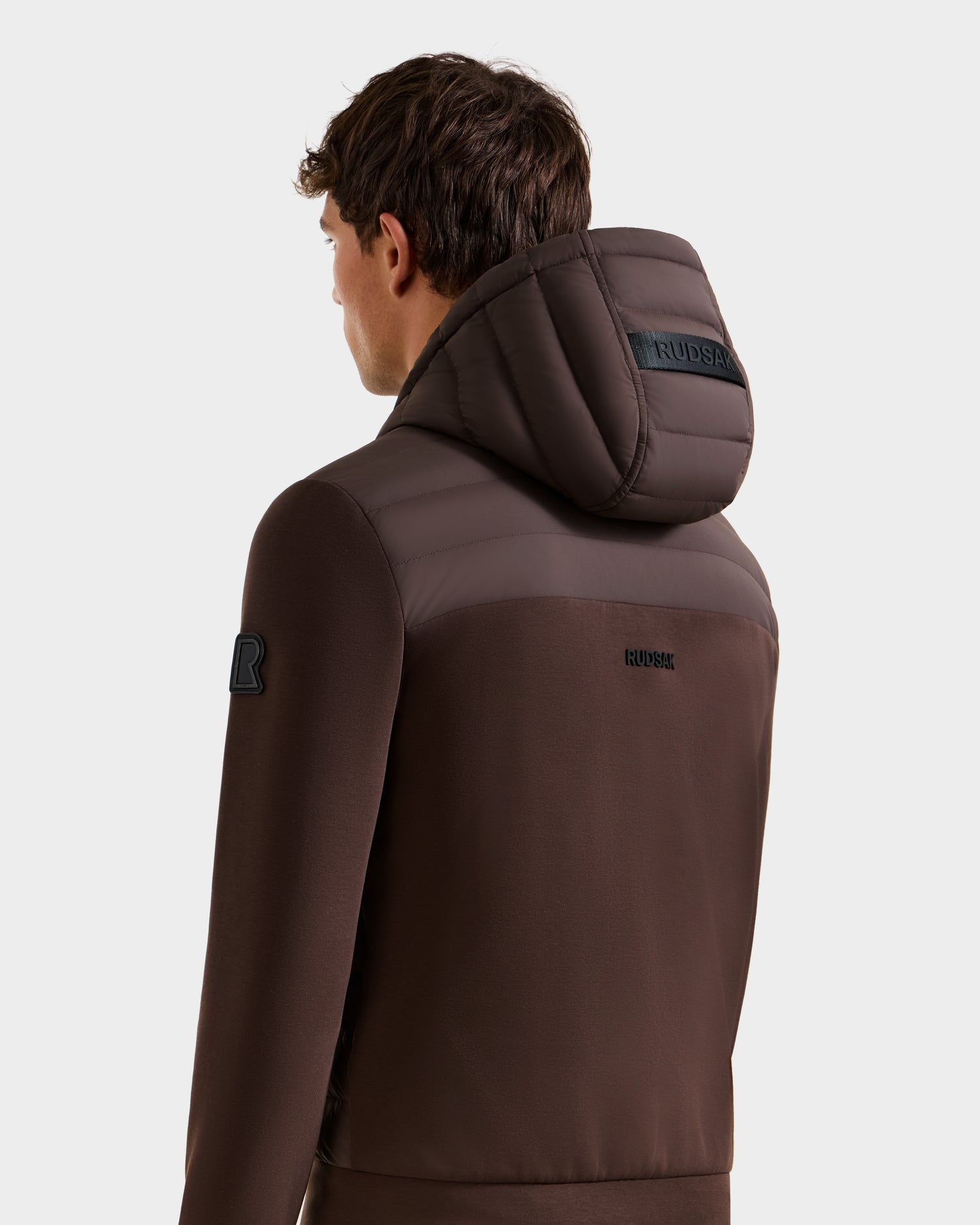 A man with short brown hair is seen from behind wearing the Rudsak DEE MAT - 6124028 Espresso men’s lightweight hybrid down jacket with quilted shoulders and raised Rudsak logo, standing out against a plain, light background.