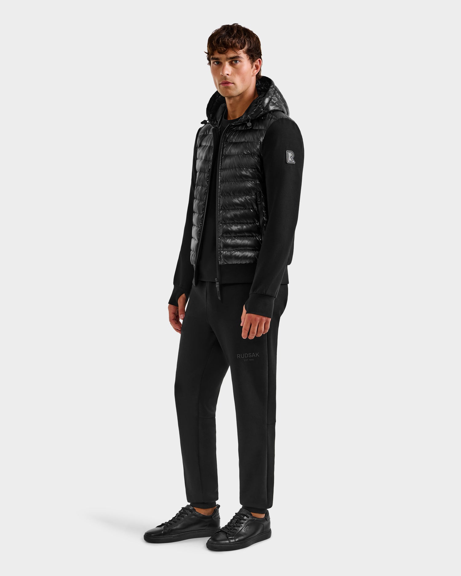 A man stands against a plain background wearing Rudsak's DEE S - 6125684 Black men’s lightweight down jacket with RDS-certified duck down, paired with black jogger pants, a zip-up hoodie, and sneakers. He faces the camera with a neutral expression.
