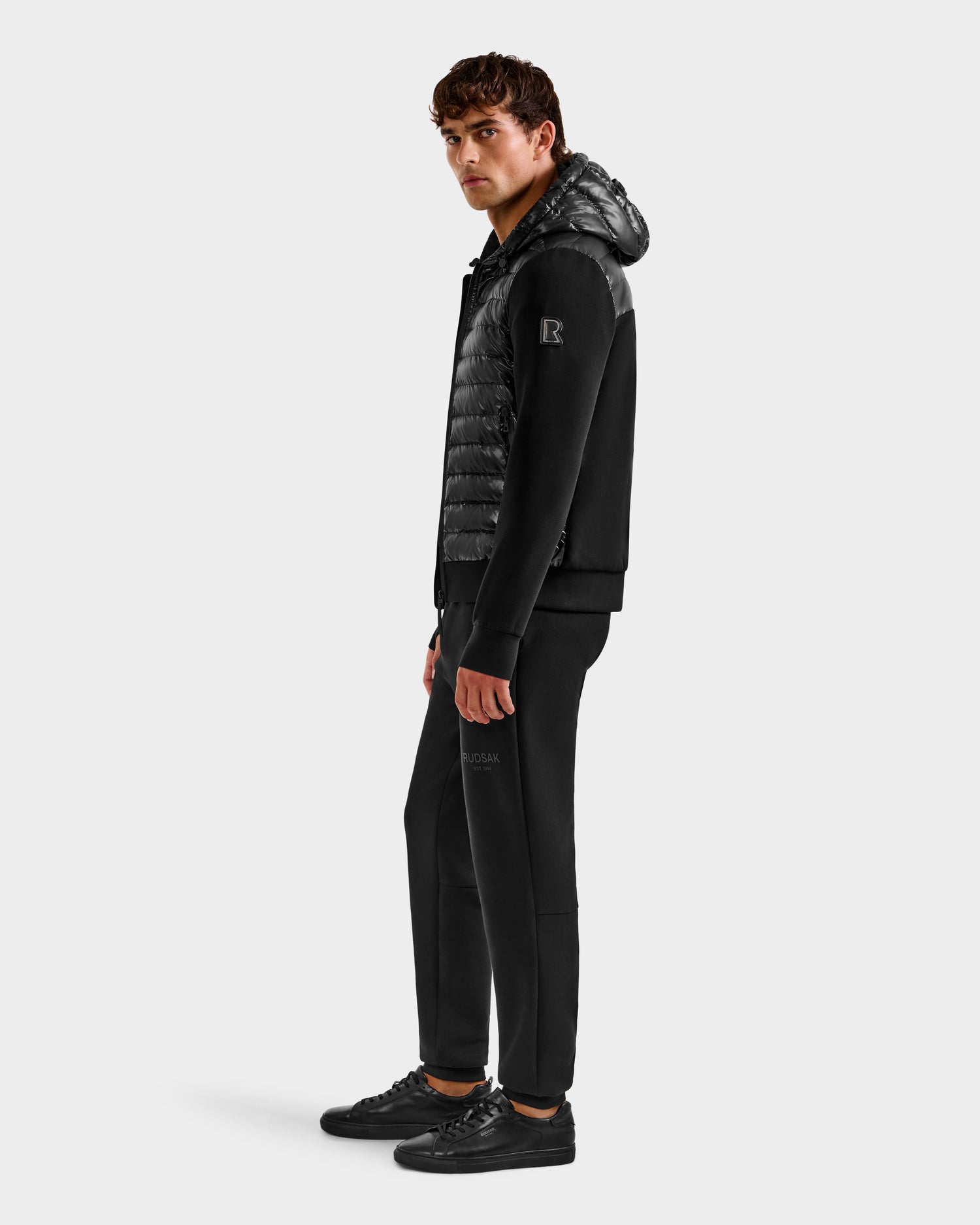 A man stands sideways wearing the Rudsak DEE S - 6125684 Black men’s lightweight down jacket with RDS-certified duck down, paired with black pants and sneakers, against a plain white background.