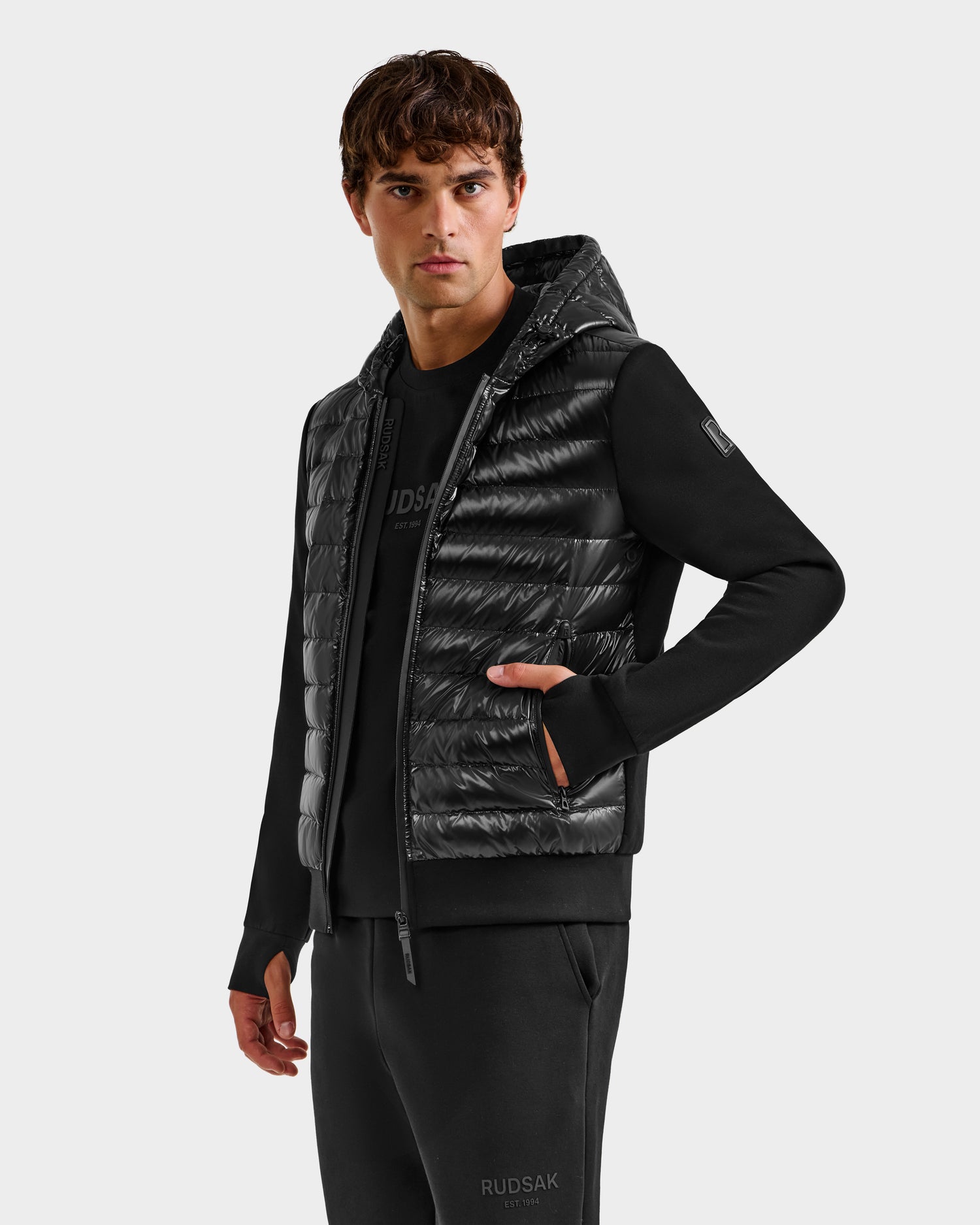 A man wears the Rudsak DEE S - 6125684 Black men's lightweight down hooded puffer vest over a black sweatshirt and pants, facing slightly left with one hand in his pocket against a plain light gray background—perfect for urban style.