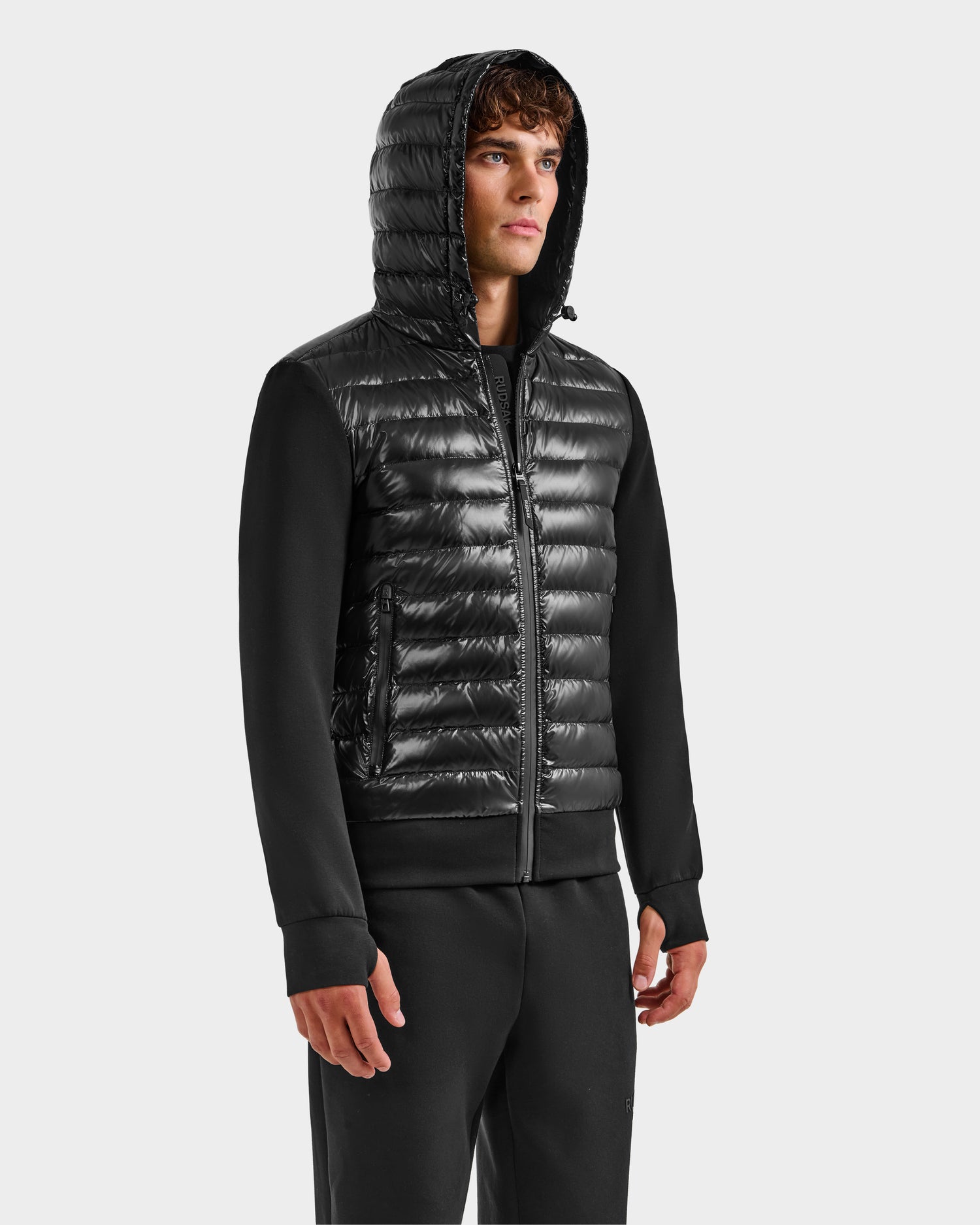 A man wears the Rudsak DEE S - 6125684 Black men’s lightweight down jacket, featuring a quilted front and smooth sleeves with RDS-certified duck down, styled with black pants and posing against a plain white background.