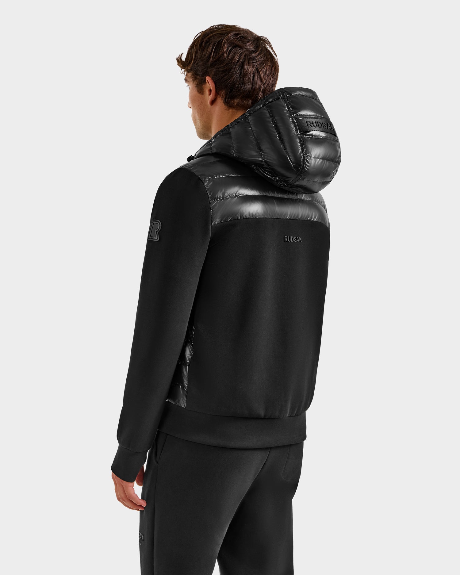 A man faces away in a Rudsak DEE S - 6125684 Black men’s lightweight down jacket with shiny quilted hood and shoulders, paired with black pants against a plain gray background.