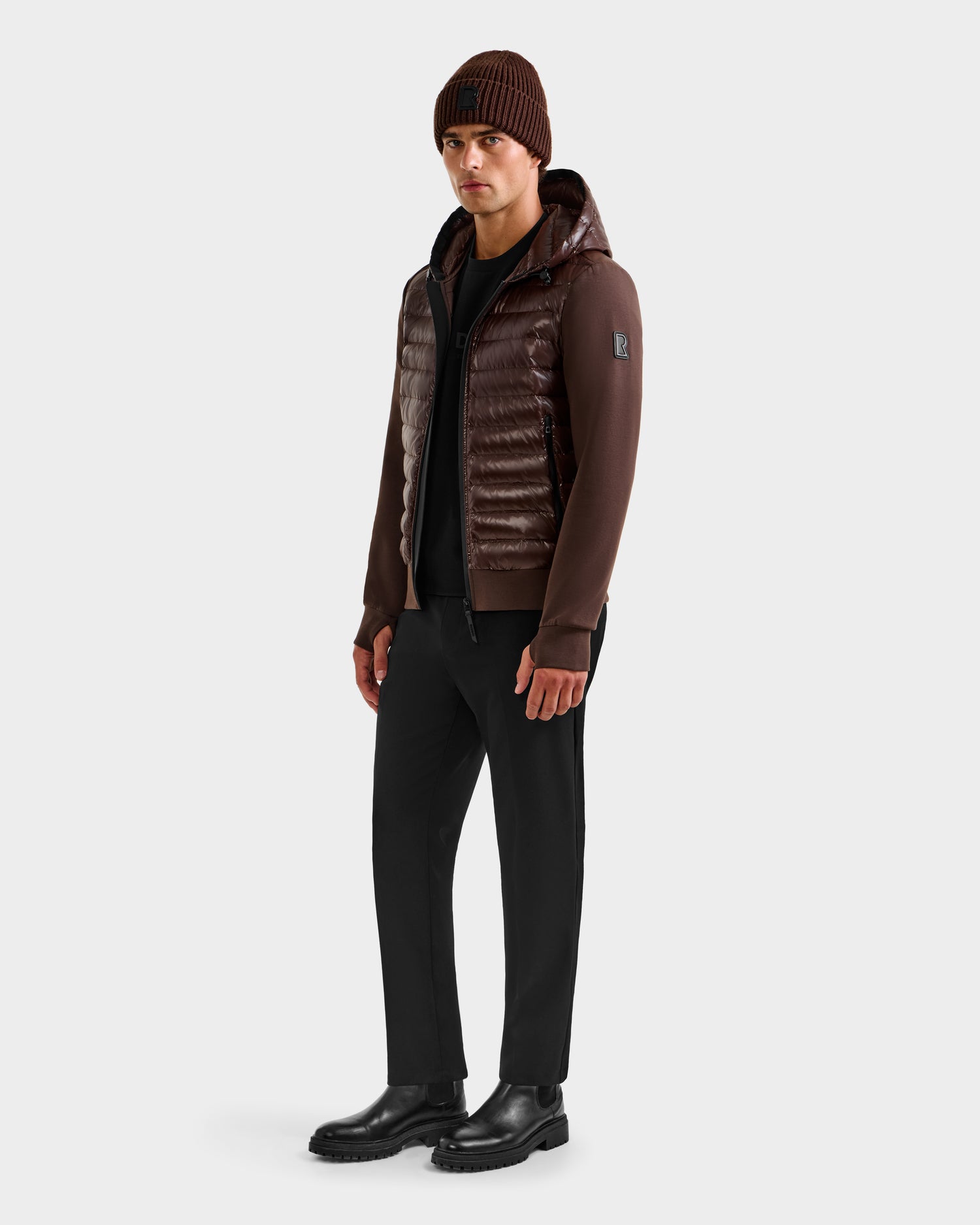 A man stands slightly sideways against a plain light background, wearing the Rudsak DEE S - 6125684 Espresso RDS-certified duck down men’s lightweight jacket—a perfect urban layer.