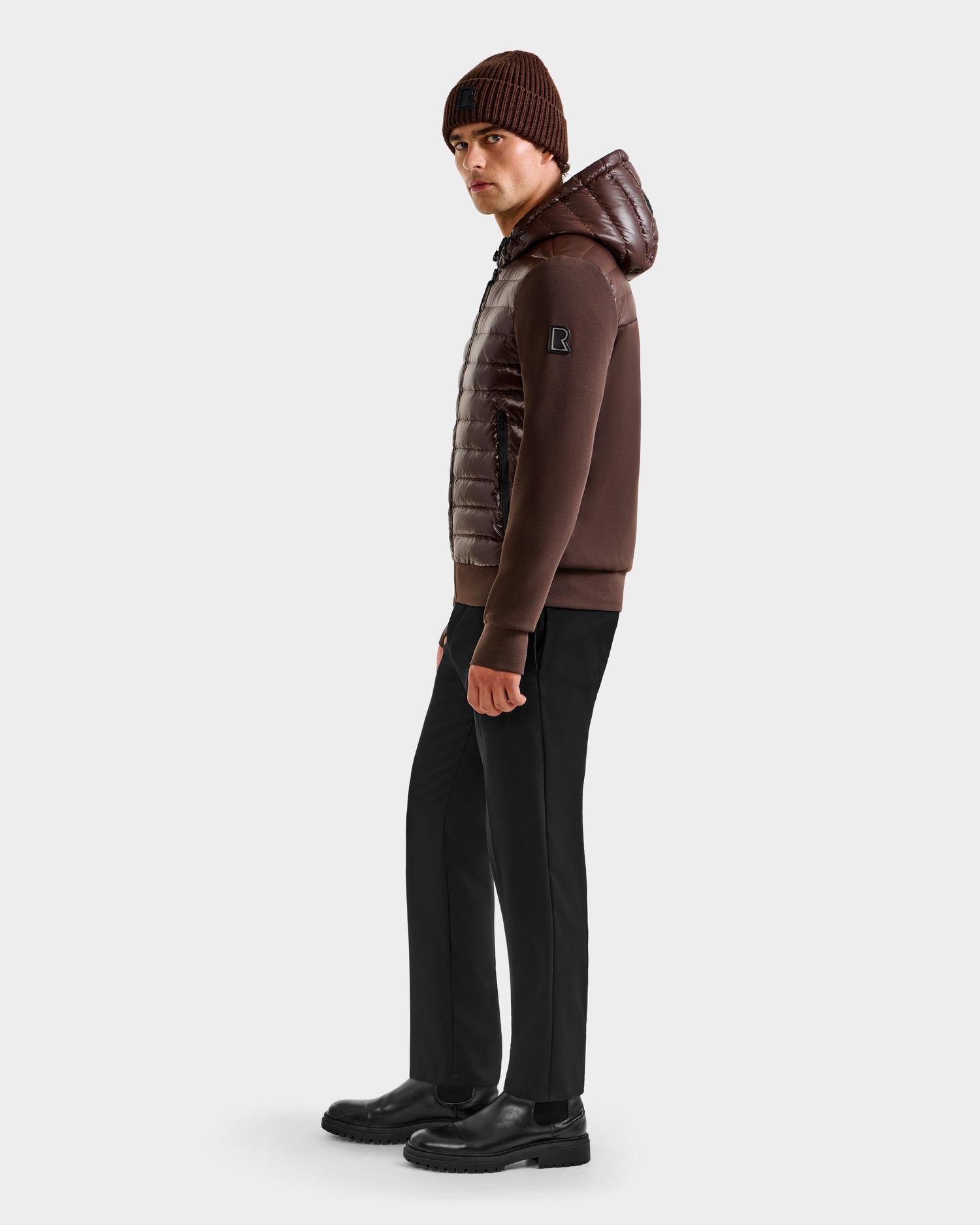 A man wearing the Rudsak DEE S - 6125684 Espresso urban layer men’s lightweight down jacket stands against a plain white background, looking to the side. He pairs it with a brown beanie, black pants, and black boots.