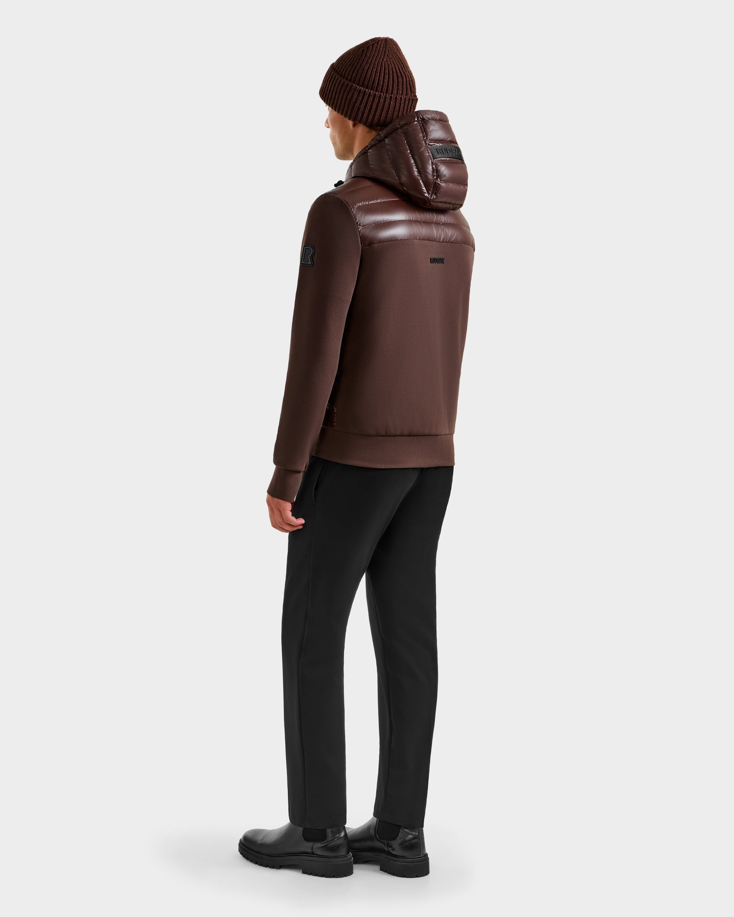 A person wearing the Rudsak DEE S - 6125684 Espresso men’s lightweight down jacket stands facing away, body slightly turned to the side against a plain light background, showcasing this ideal urban layering piece.