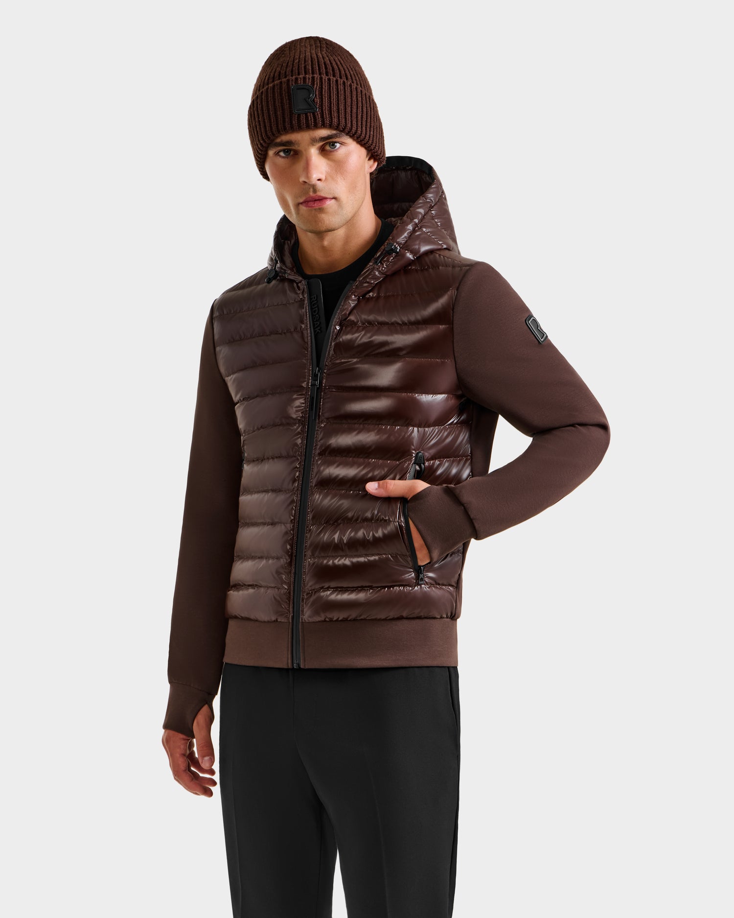 A man wearing the Rudsak DEE S - 6125684 Espresso brown hooded puffer jacket with knit sleeves, a brown beanie, and black pants stands against a white background, showcasing this stylish men’s lightweight down layer.