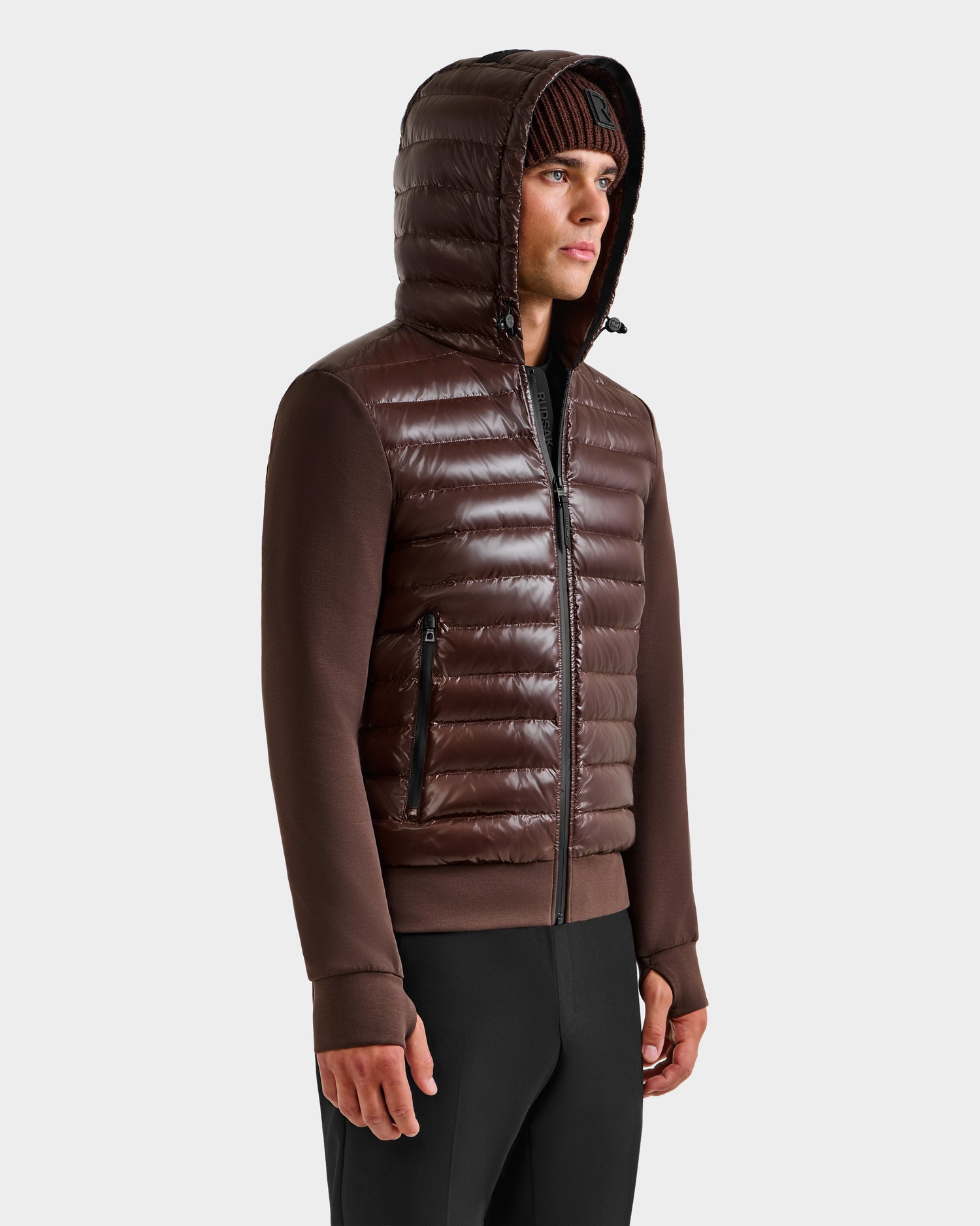 A man in the Rudsak DEE S - 6125684 Espresso lightweight down jacket with knit sleeves, a brown knit beanie, and black pants stands against a light gray background—an urban layer for cool weather.