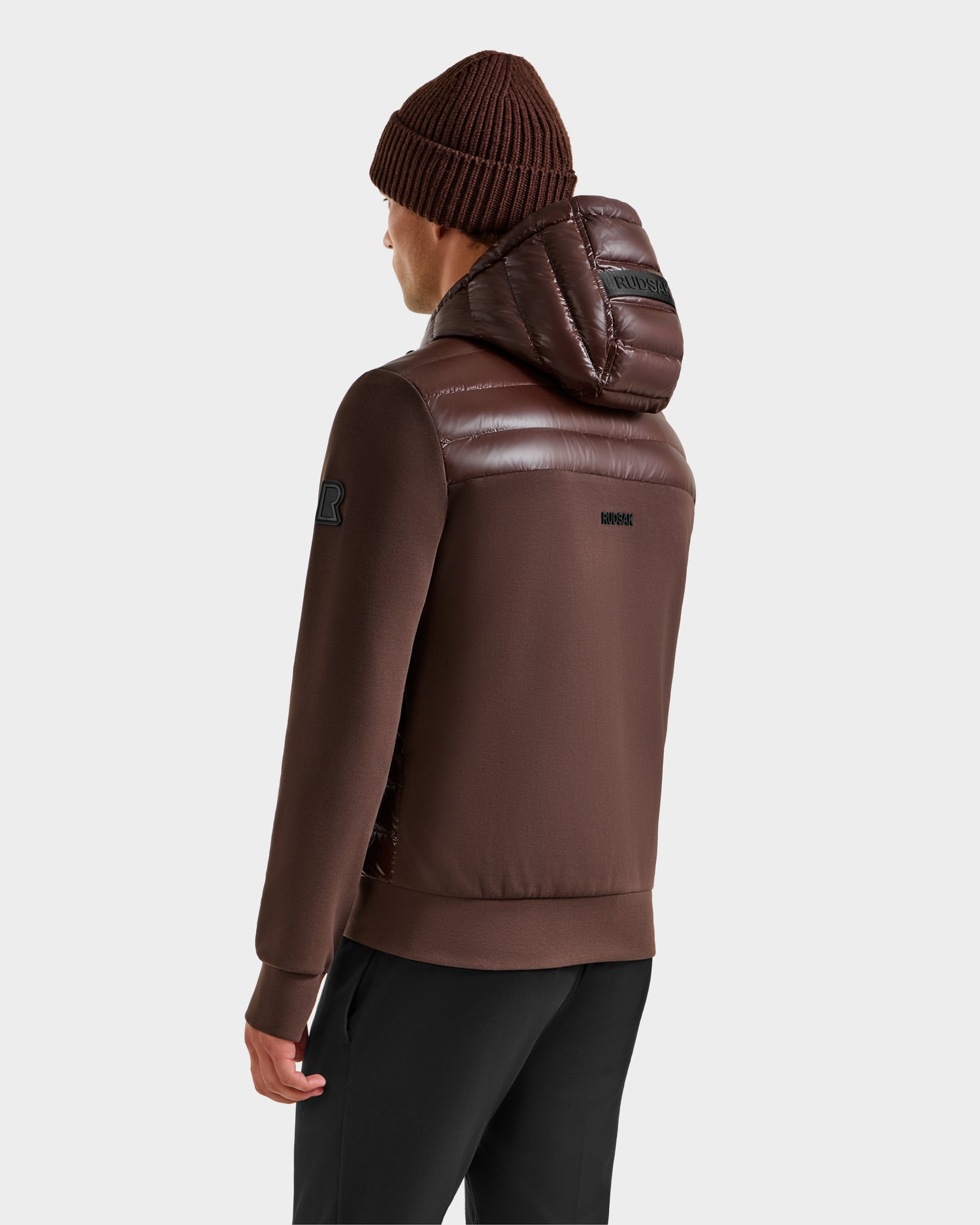 A person in a brown beanie and an espresso DEE S - 6125684 men's lightweight down jacket by Rudsak, featuring quilted hood and shoulder sections plus an upper sleeve patch, stands facing away.
