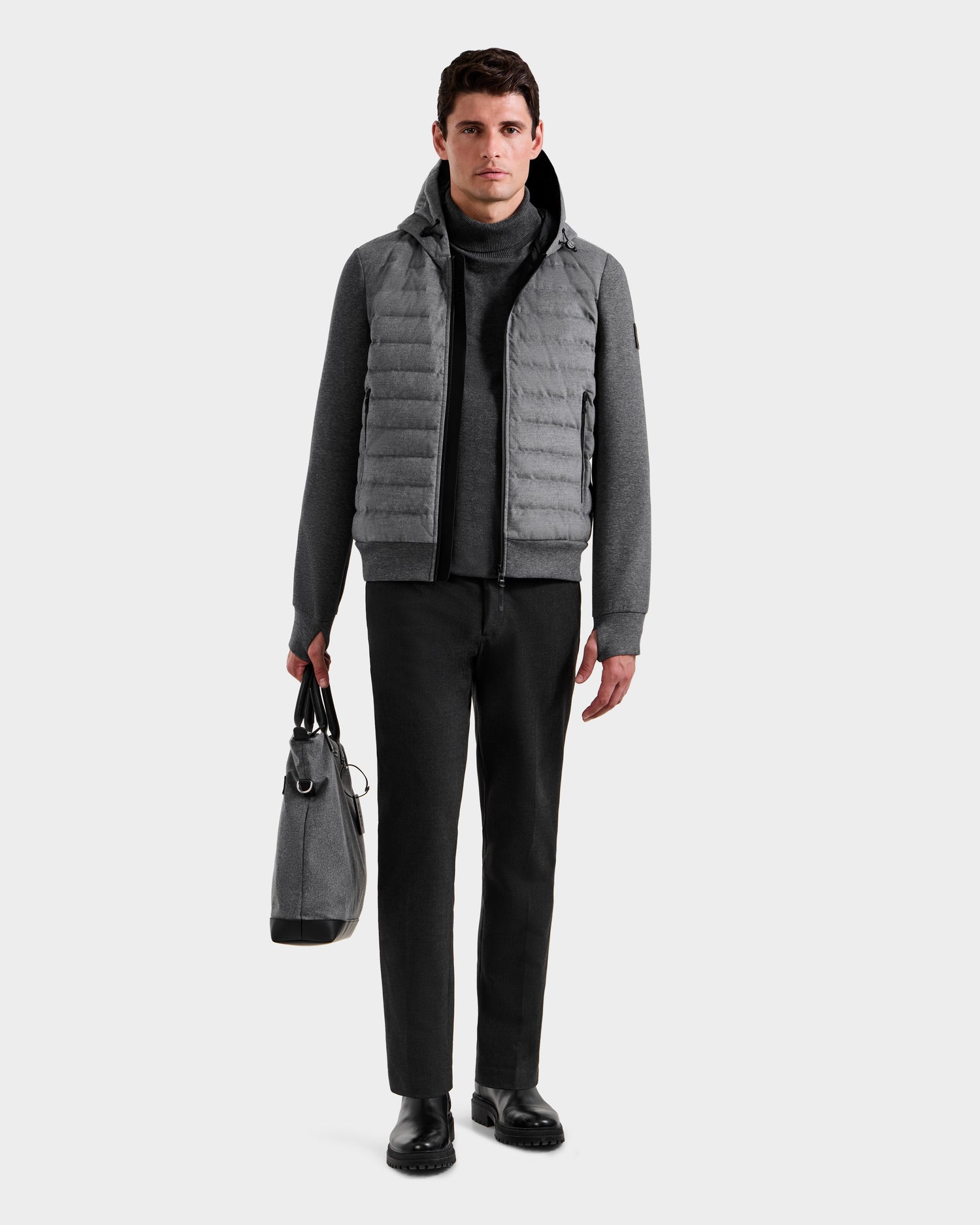 A man faces forward, wearing the Rudsak DEE W - 6125570 Heather Charcoal men’s quilted down jacket over a turtleneck, black pants, and black shoes. He holds a grey bag in his left hand against a plain light background.