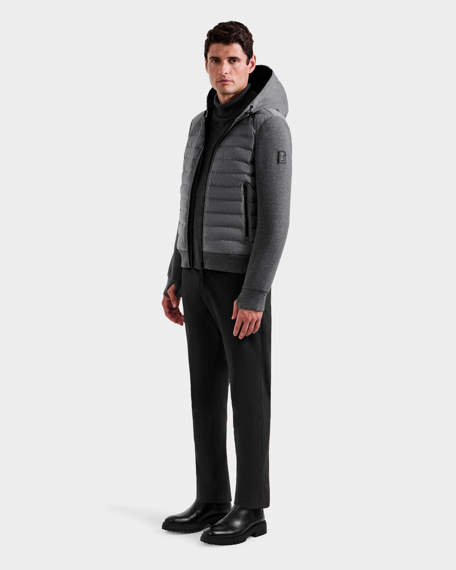 A man stands slightly turned, wearing the Rudsak DEE W - 6125570 Heather Charcoal men's quilted down jacket, paired with dark pants and black boots, against a plain white background.