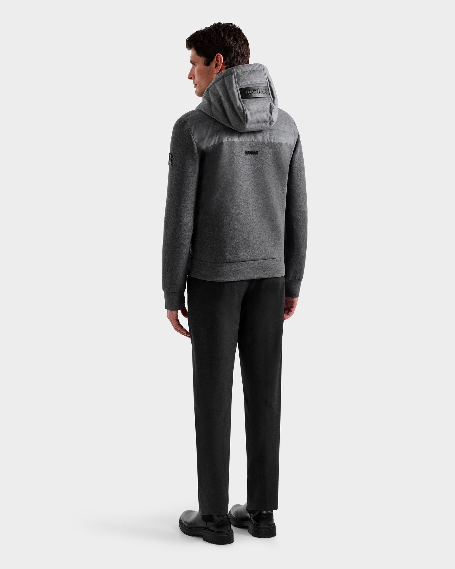 A man faces away, wearing the Rudsak DEE W - 6125570 Heather Charcoal men's quilted down jacket, paired with black pants and shoes against a plain white background.