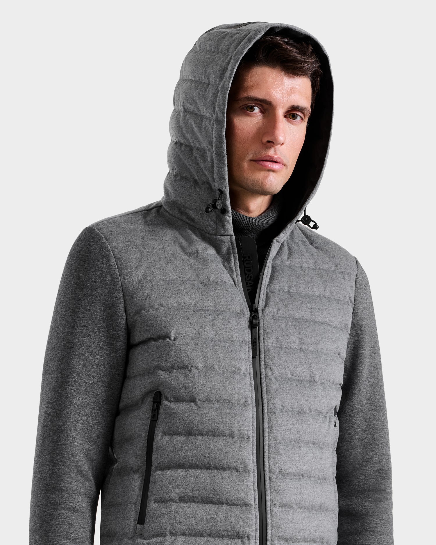 A man wears the Rudsak DEE W - 6125570 Heather Charcoal jacket, a gray, lightweight insulated piece with a quilted design, hood, zipper, and zippered pockets, standing against a plain light background.