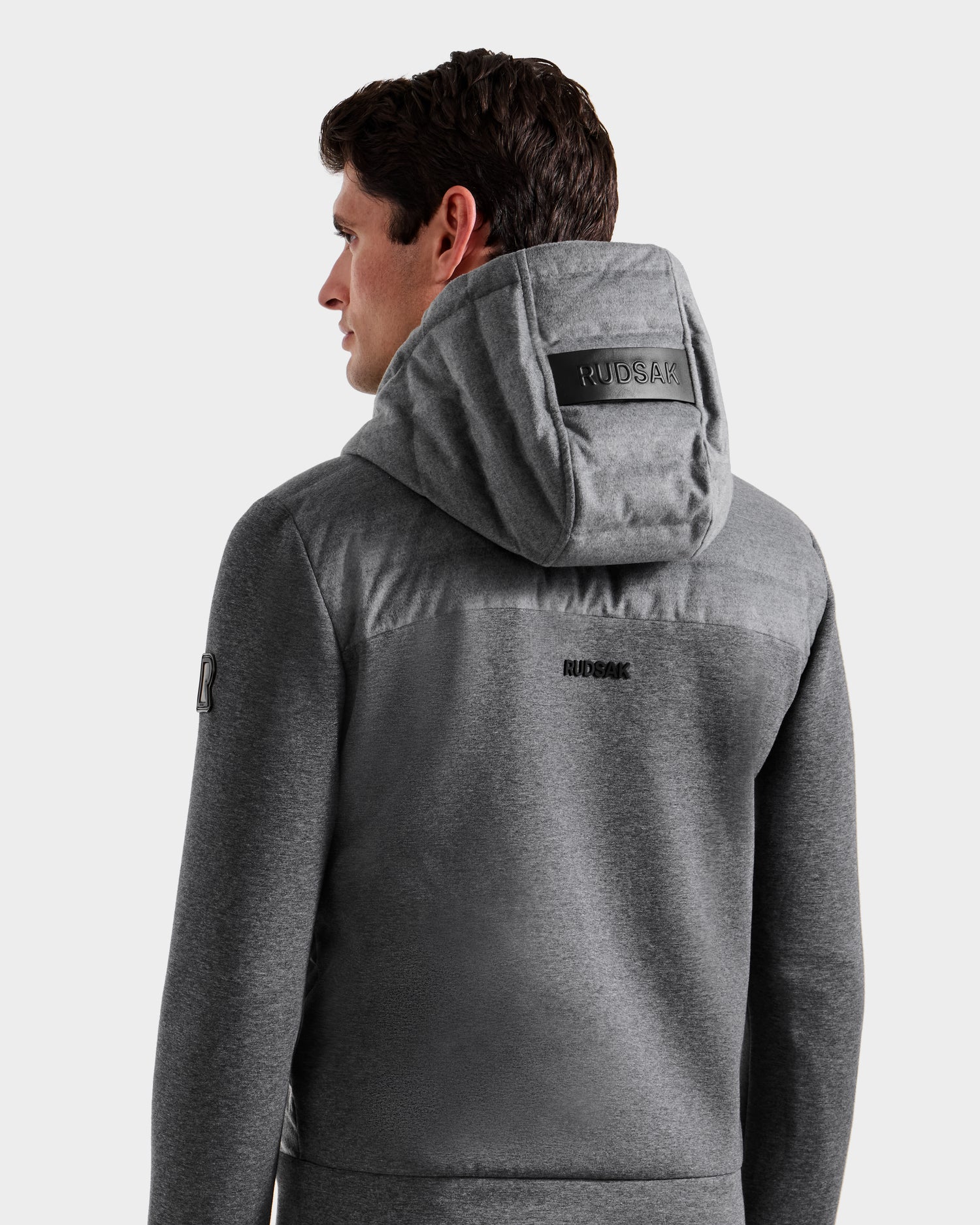 A man with short brown hair faces away from the camera, wearing a Rudsak DEE W - 6125570 Heather Charcoal lightweight insulated jacket with “RUDSAK” branding on the back, set against a plain light background.