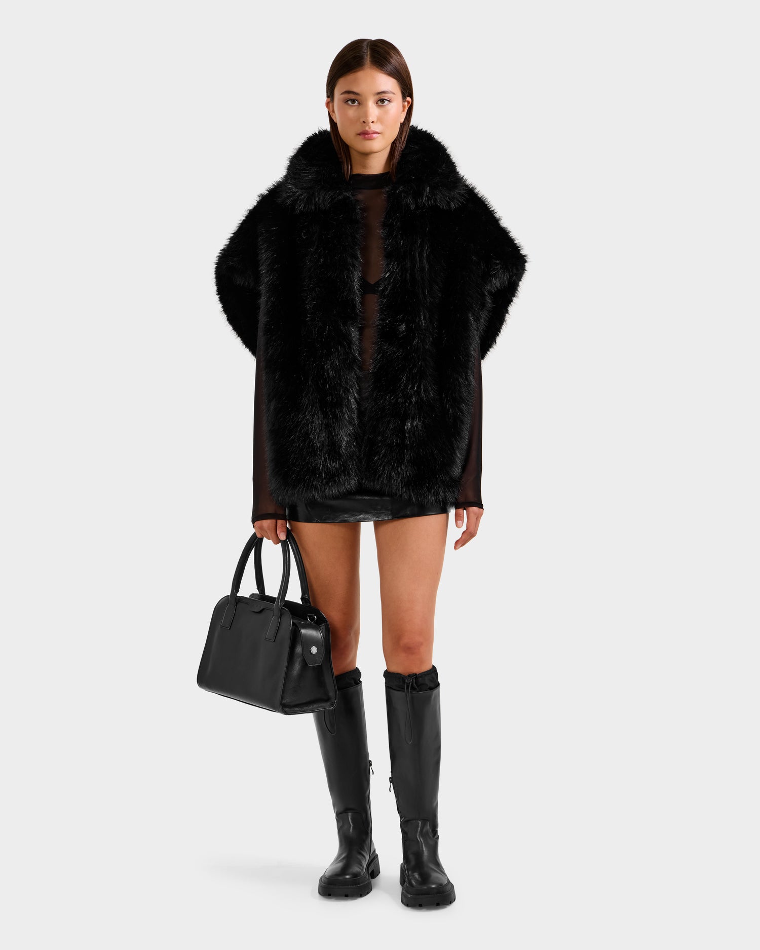 A woman faces forward in the Rudsak DELFINA - 8525662 Black faux fur cape with an oversized silhouette, styled with a sheer black top, mini skirt, knee-high boots, and a black handbag—a chic winter outerwear look.