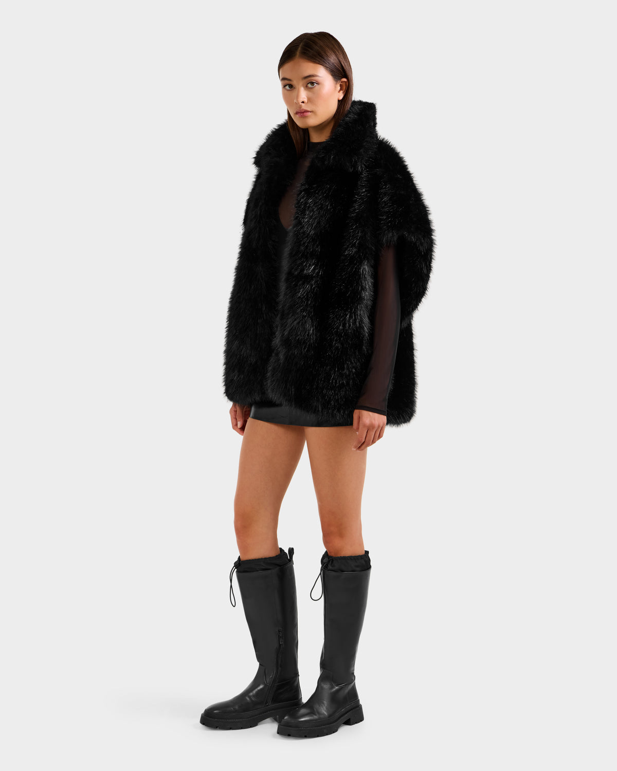A woman wears the Rudsak DELFINA - 8525662 Black faux fur vest with an oversized silhouette, paired with a black mini skirt, sheer top, and knee-high boots against a plain light background.