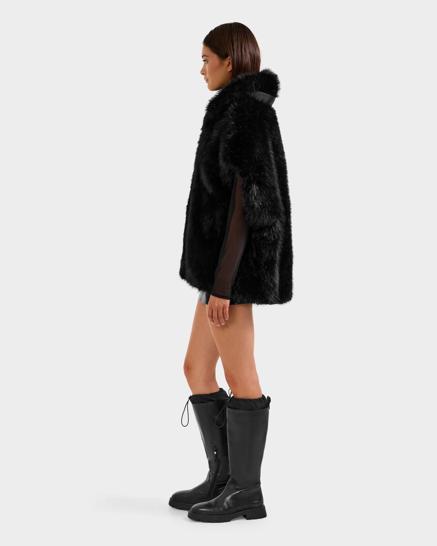 A woman stands in profile, showcasing the Rudsak DELFINA - 8525662 Black faux fur coat with an oversized silhouette, paired with black knee-high boots and a dark outfit, set against a plain light background.