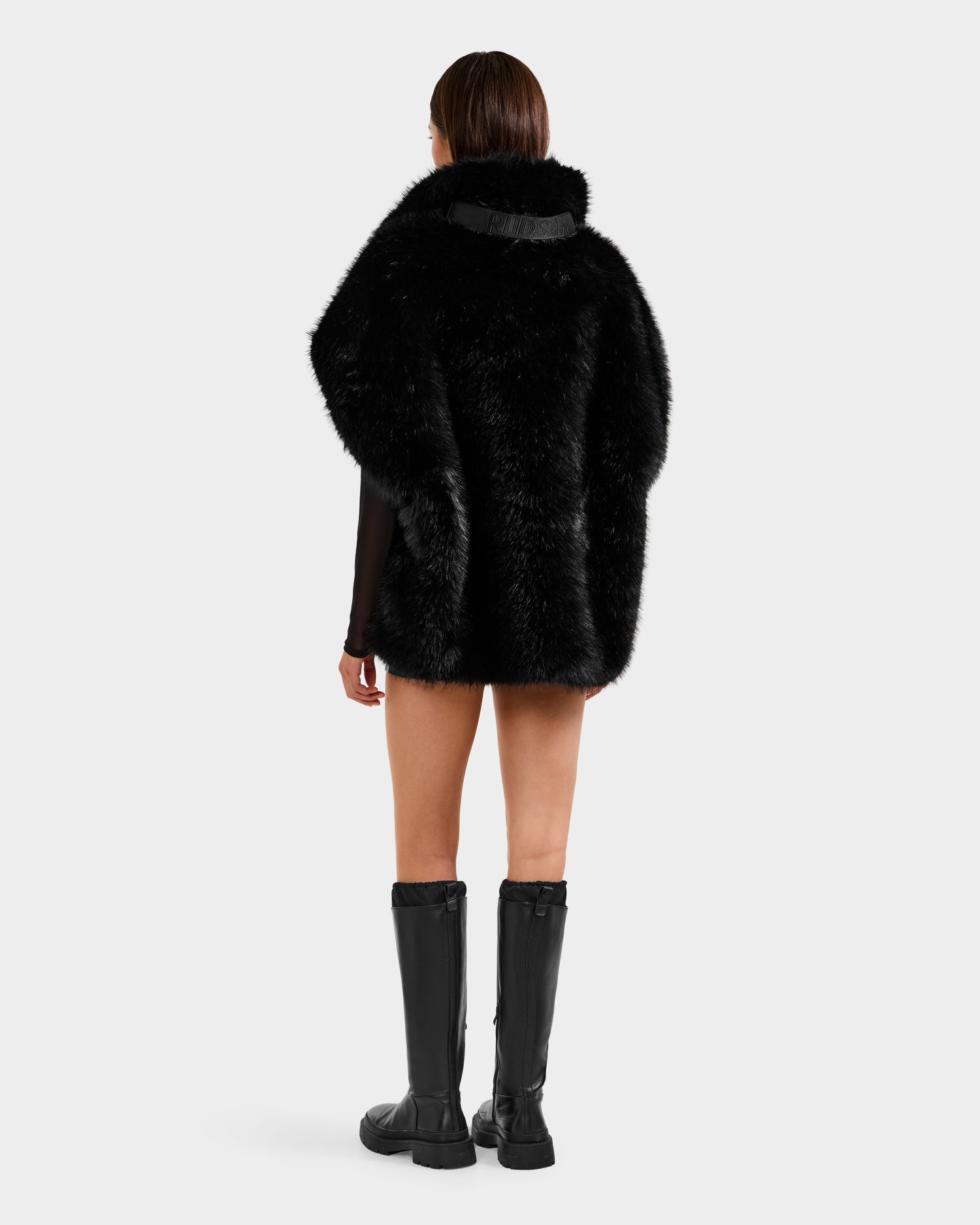 A person with short, straight brown hair faces away, wearing the Rudsak DELFINA - 8525662 Black faux fur winter coat and knee-high black boots, set against a plain white background.