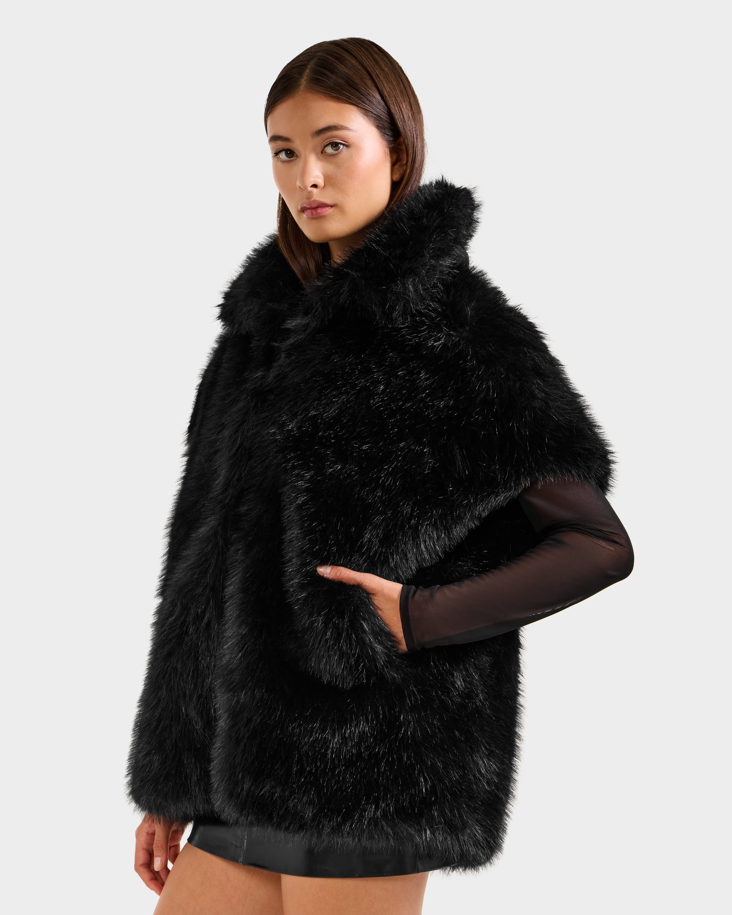 A young woman wears the Rudsak DELFINA - 8525662 Black faux fur coat with a high collar over a sheer top and short skirt, showcasing chic winter outerwear against a plain light gray background.