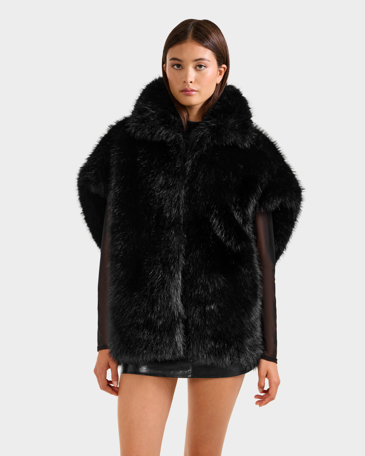 A woman faces forward wearing the Rudsak DELFINA - 8525662 Black faux fur coat with a large collar and oversized silhouette, paired with a short black mini skirt against a plain light gray background.