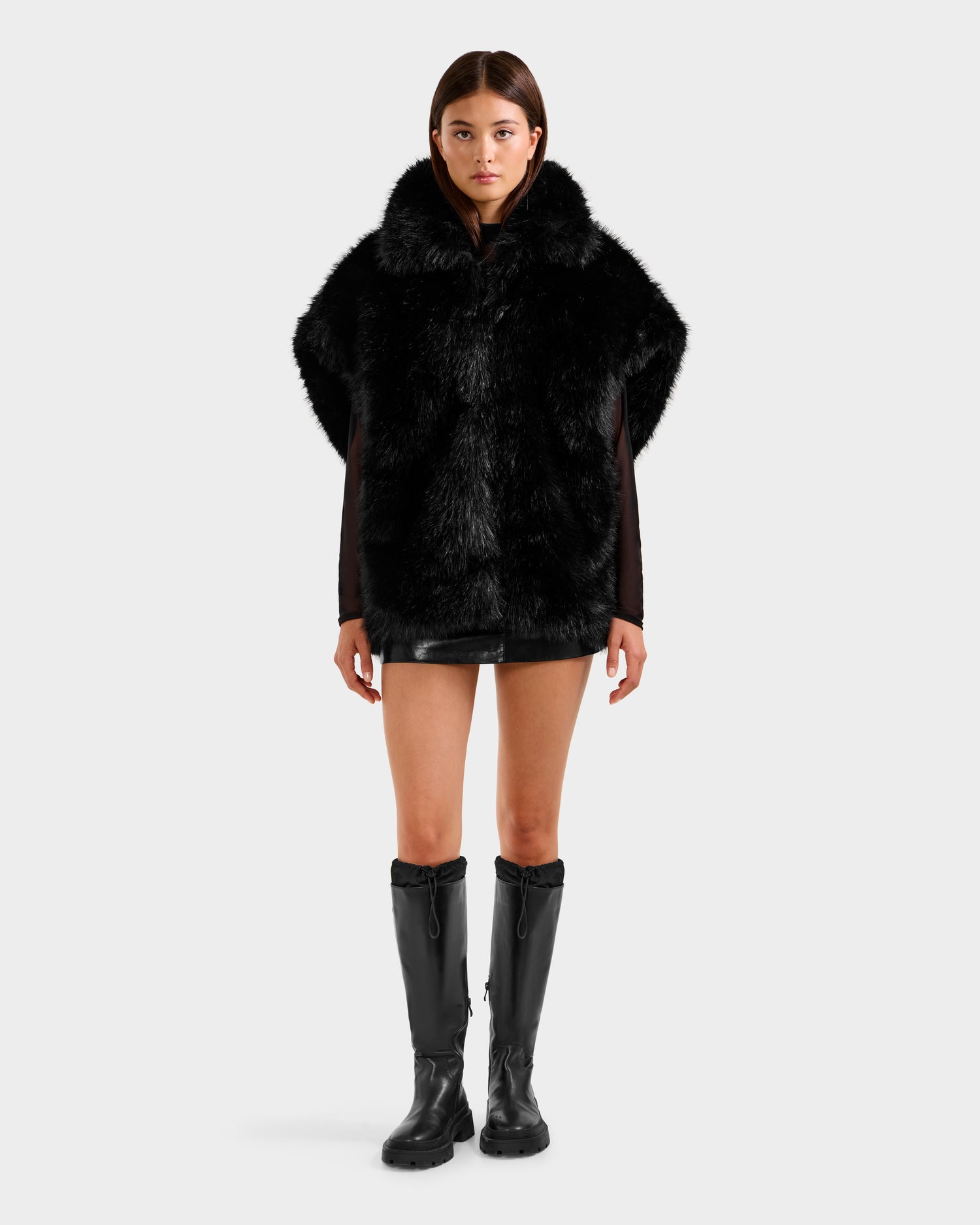A person faces forward in the Rudsak DELFINA - 8525662 black faux fur cape with an oversized silhouette, paired with a black mini skirt and knee-high boots, against a plain light background.