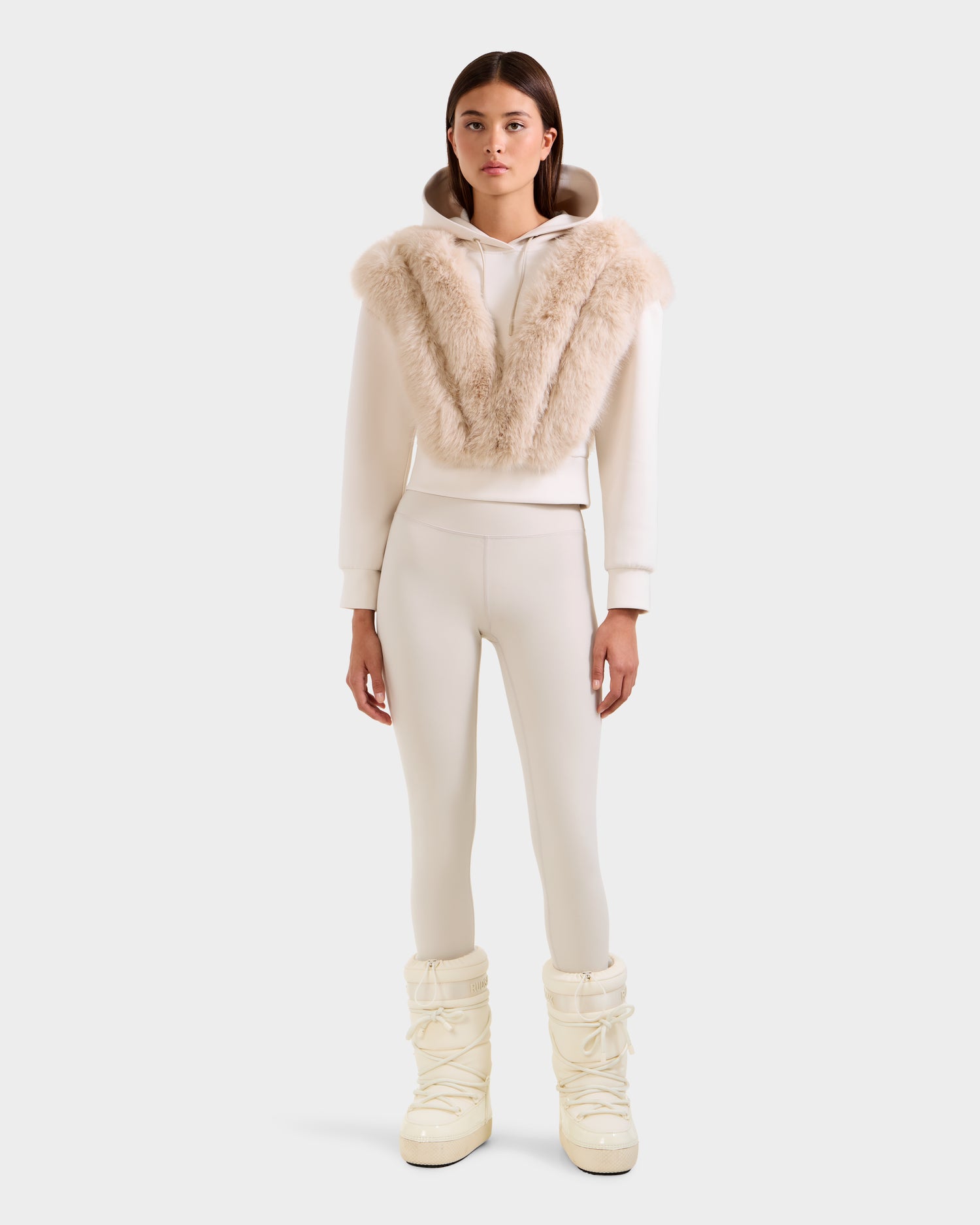 A woman faces forward wearing the Rudsak DELLA - 8525668 Vanilla off-white faux fur hoodie with a large beige collar, matching fitted pants, and cream snow boots for an effortlessly chic après-ski look against a plain light background.