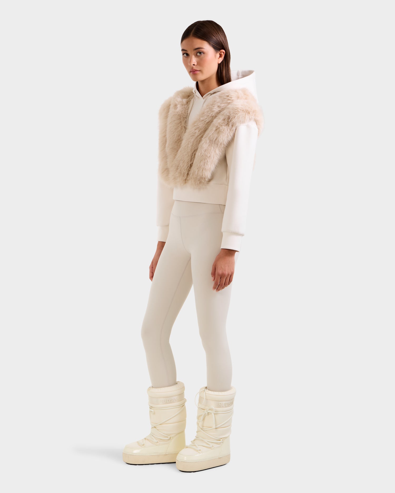 A woman wears the Rudsak DELLA - 8525668 Vanilla cropped hoodie, paired with a light beige faux fur scarf, cream leggings, and off-white Moon Boot-style snow boots, standing against a plain light gray background.
