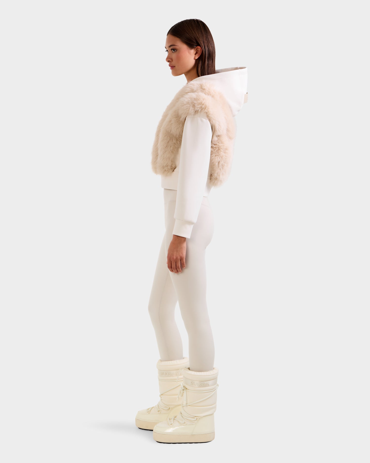 A woman in profile wears Rudsak’s DELLA - 8525668 Vanilla cropped women’s hoodie layered with a beige faux fur vest, paired with white leggings and cream snow boots against a plain light background.