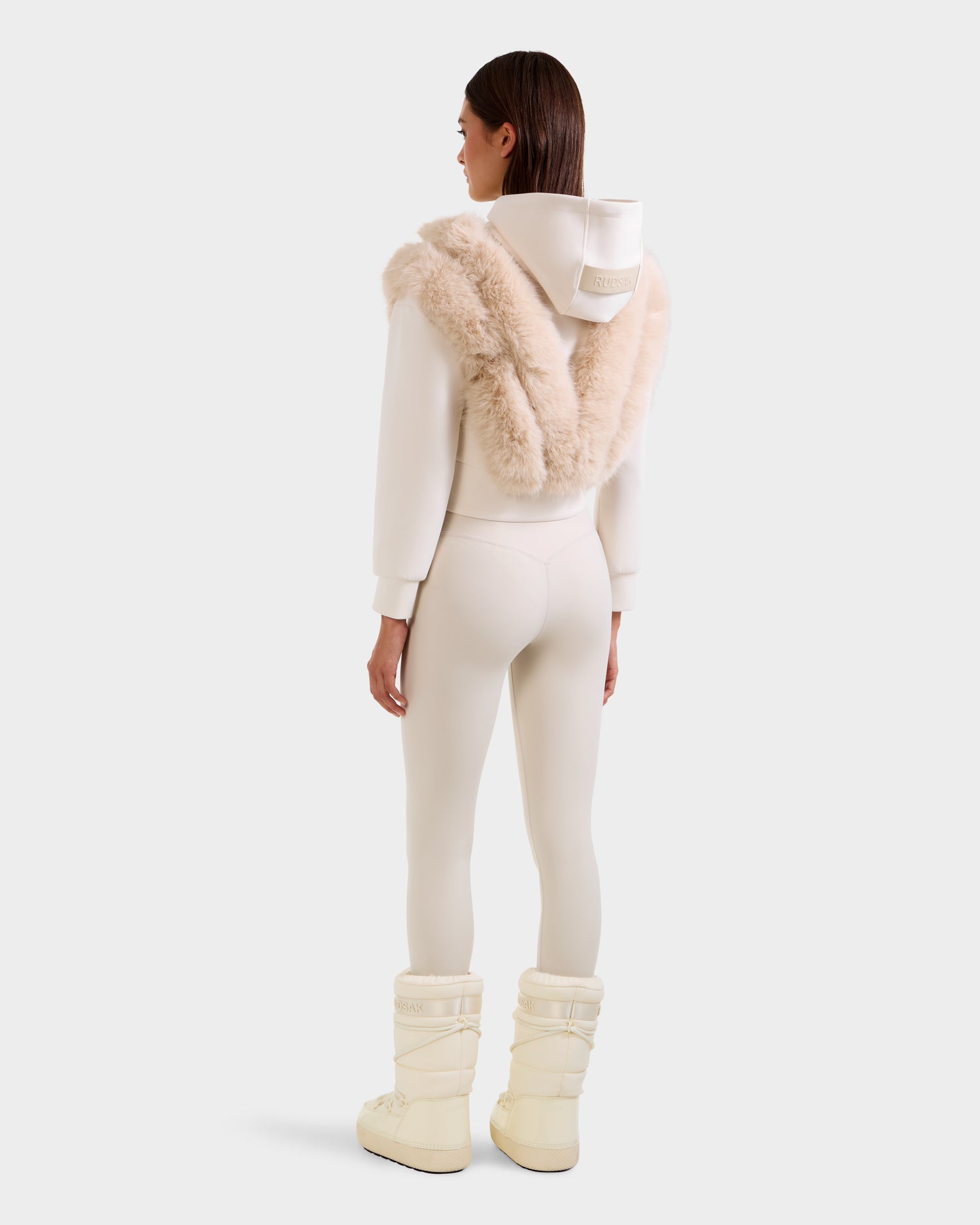 A woman stands facing away in a Rudsak DELLA - 8525668 Vanilla off-white faux fur hoodie, beige scarf, matching fitted pants, and cream winter boots against a plain light grey background.