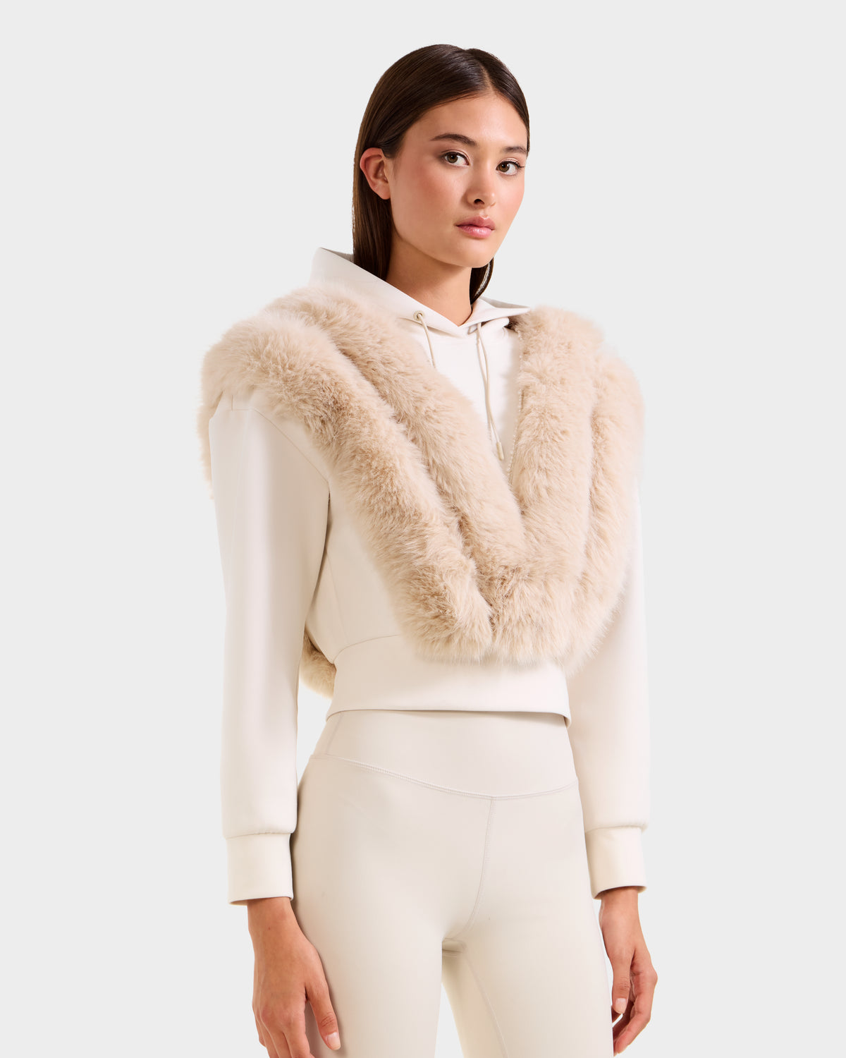 A woman wearing the Rudsak DELLA - 8525668 Vanilla cream après-ski pullover with a faux fur shawl draped over her shoulders stands against a plain white background.