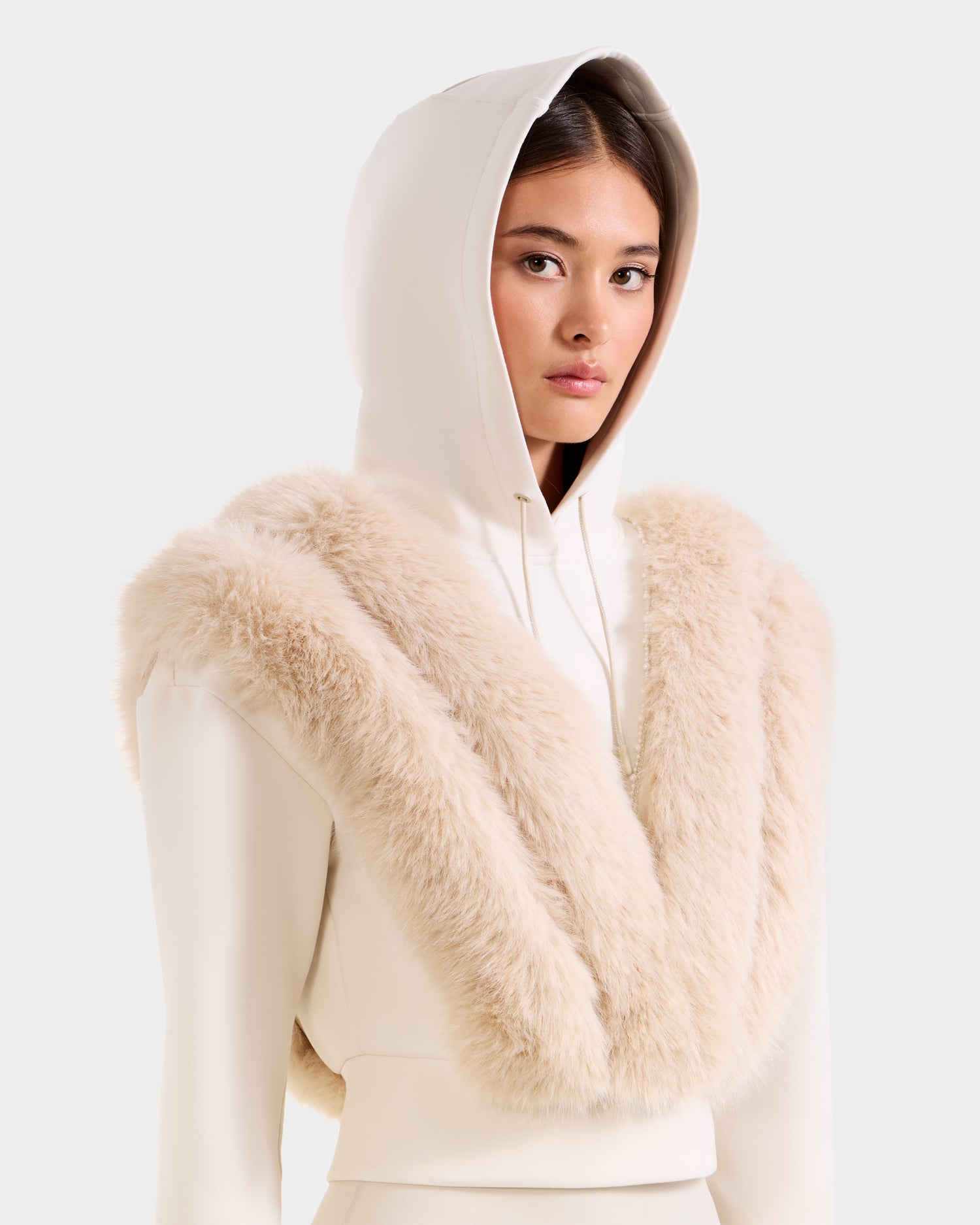 A woman stands confidently against a plain light background, wearing the Rudsak DELLA - 8525668 Vanilla après-ski pullover and a plush, light beige faux fur wrap draped over her shoulders.