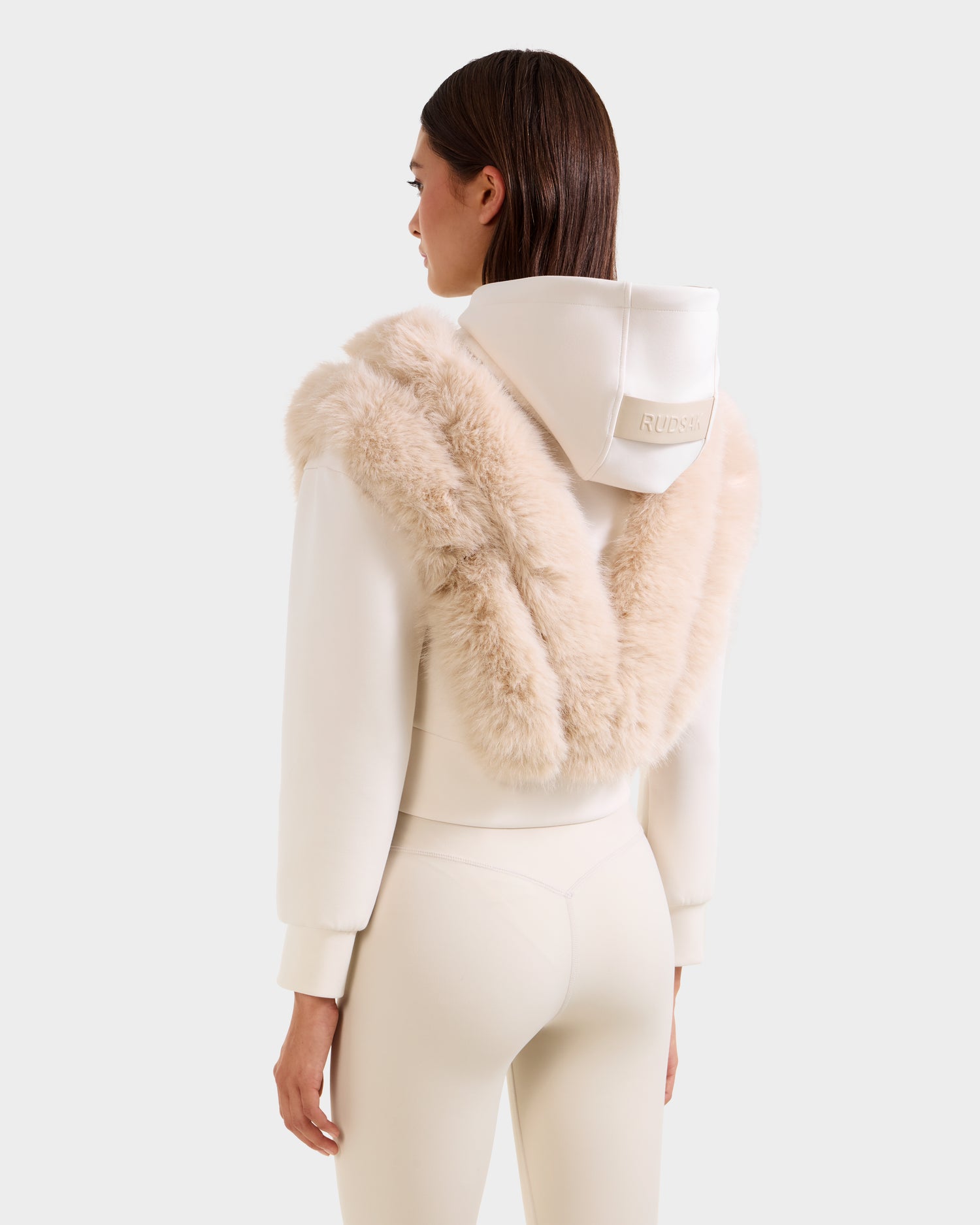 A woman models the Rudsak DELLA - 8525668 Vanilla cream faux fur hoodie with a large fluffy trim, paired with matching high-waisted leggings. The cozy après-ski pullover is shown from the back against a plain backdrop.