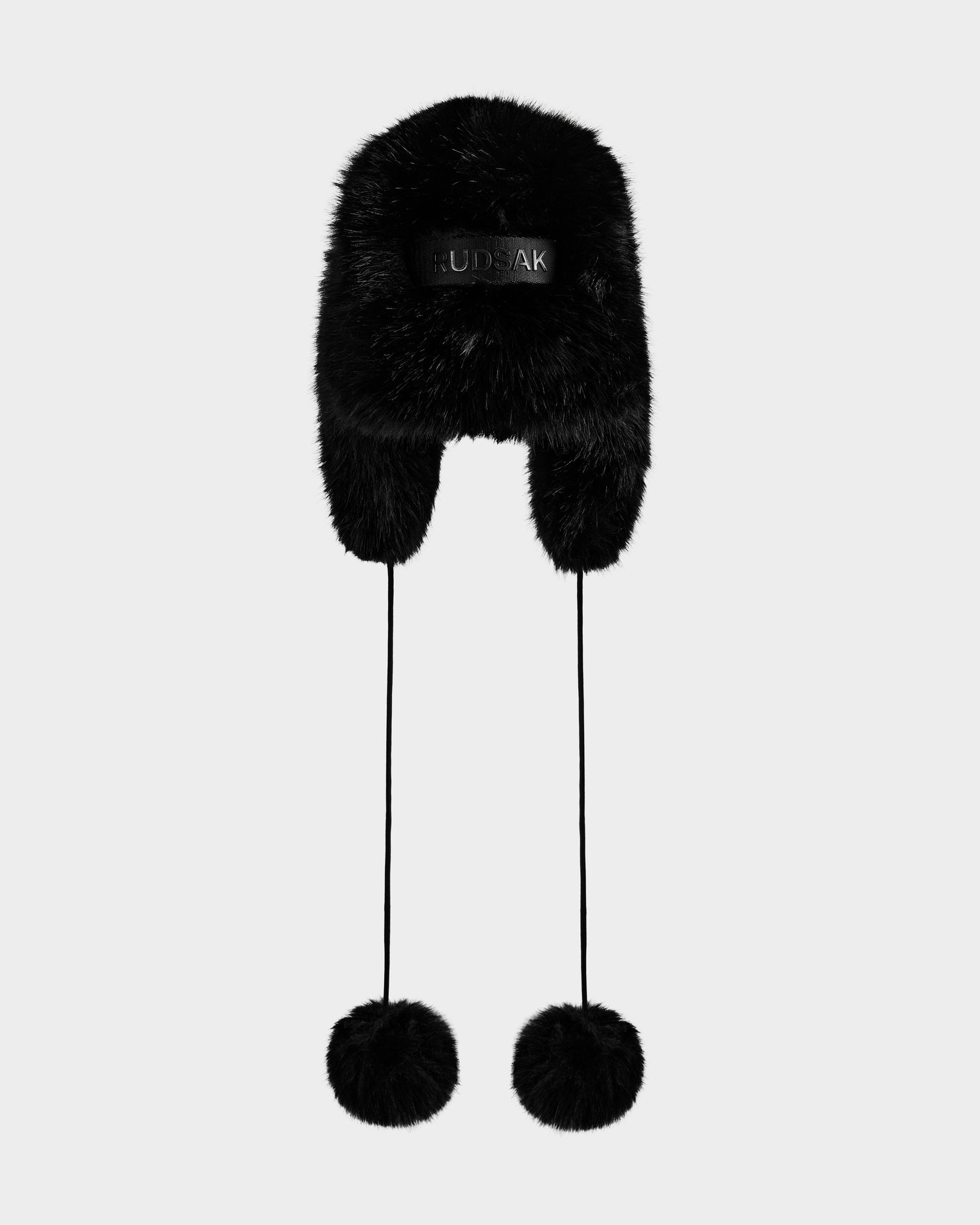 The Rudsak DEMI - L425539 Black faux-fur aviator hat for women features ear flaps, long strings with fluffy pom-poms, and is shown on a light gray background—a must-have winter accessory for cold-weather style.