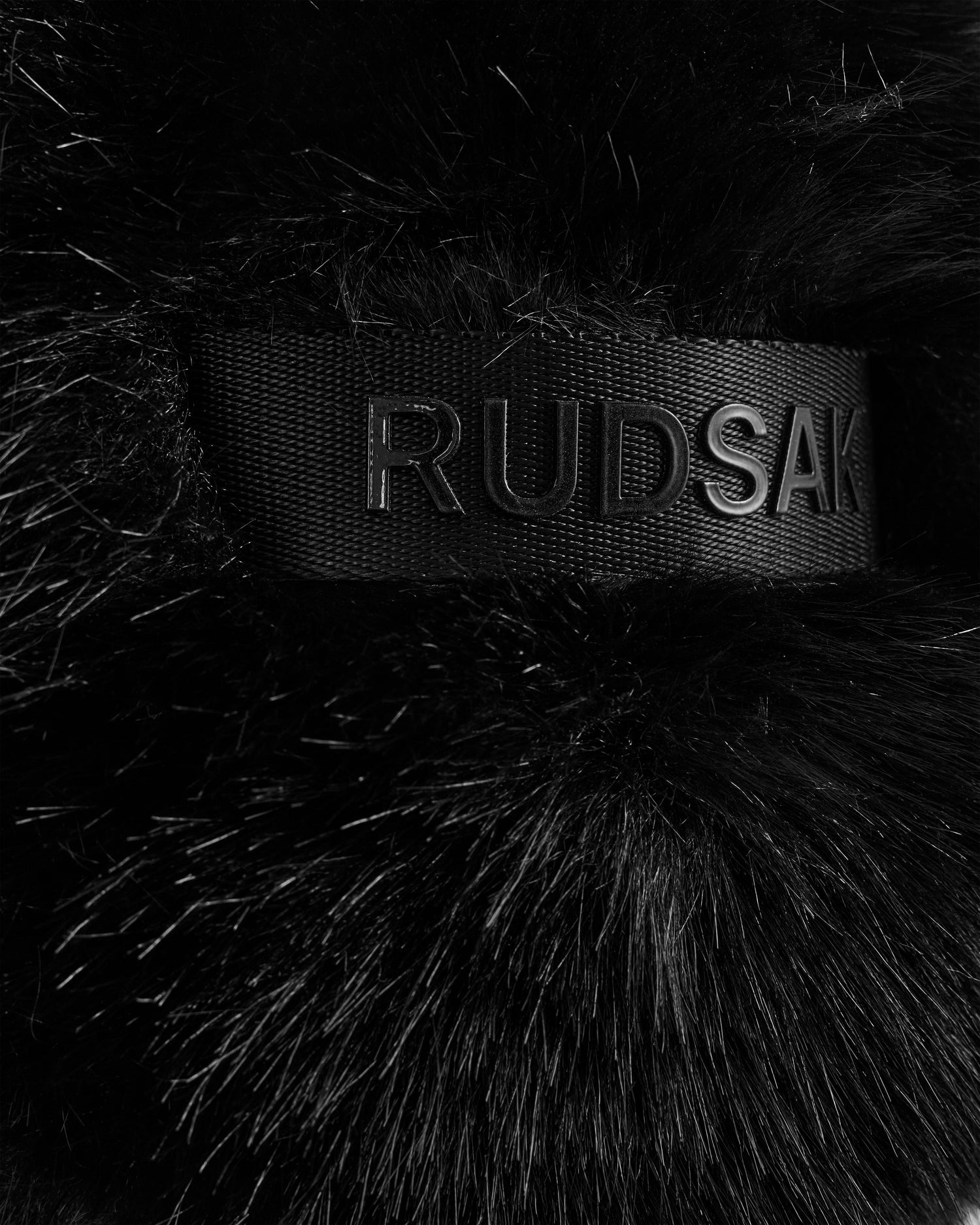 Close-up of the DEMI - L425539 Black aviator hat by Rudsak, crafted from soft black faux fur and featuring a black ribbon embossed with "RUDSAK" in bold, uppercase letters.