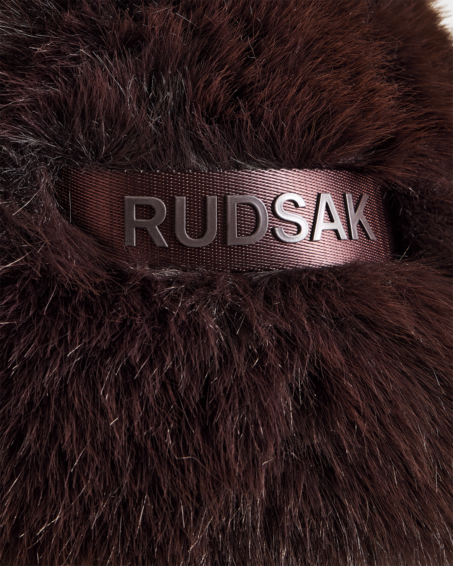 Close-up of espresso brown, furry fabric from the DEMI - L425539 women’s faux-fur aviator hat by Rudsak, featuring a brown strap with metallic embossed RUDSAK text.