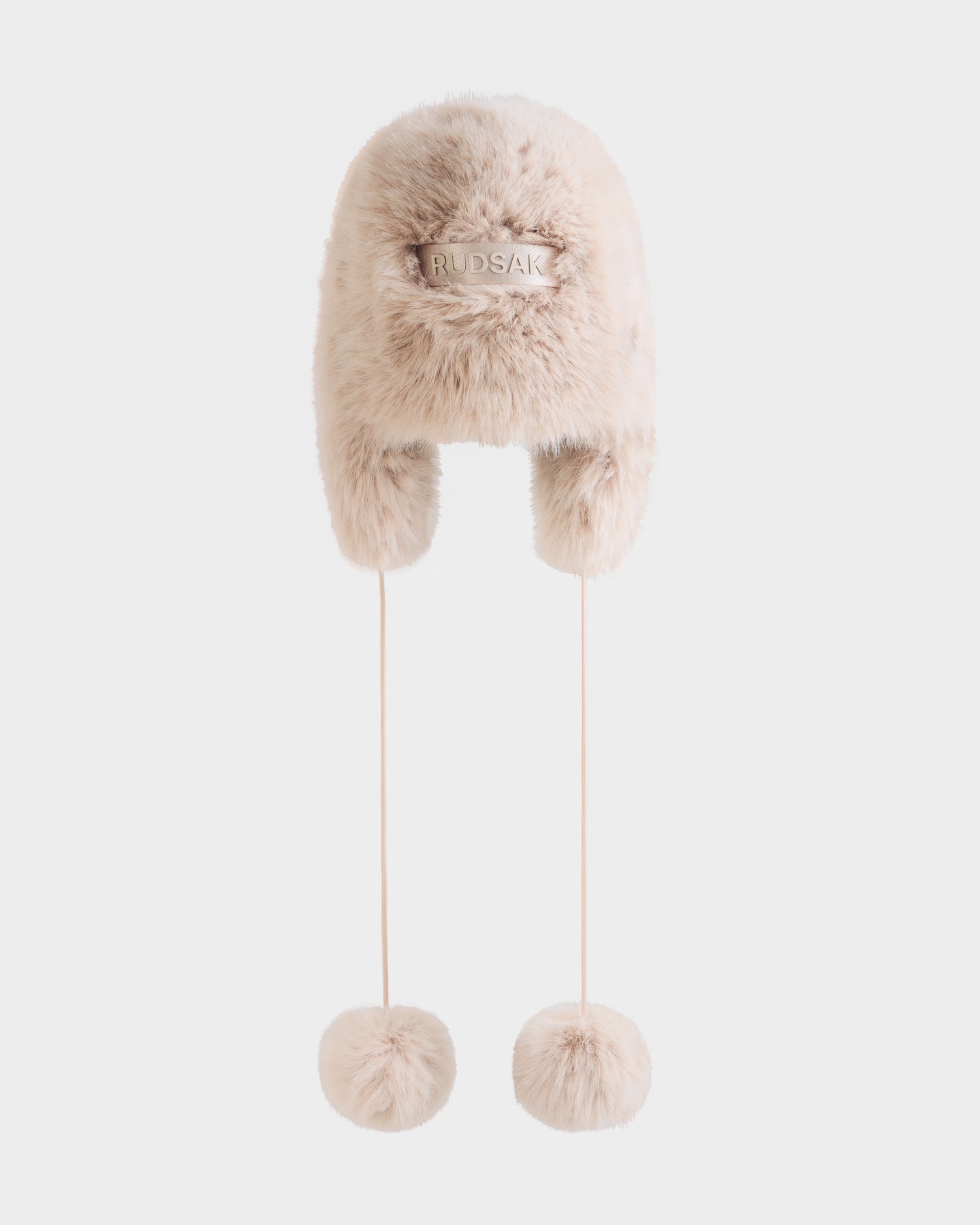 The DEMI - L425539 Vanilla by Rudsak is a women’s light beige faux-fur aviator hat with ear flaps, long strings topped with large fur pom-poms, and a branded front label—ideal for staying cozy in winter.