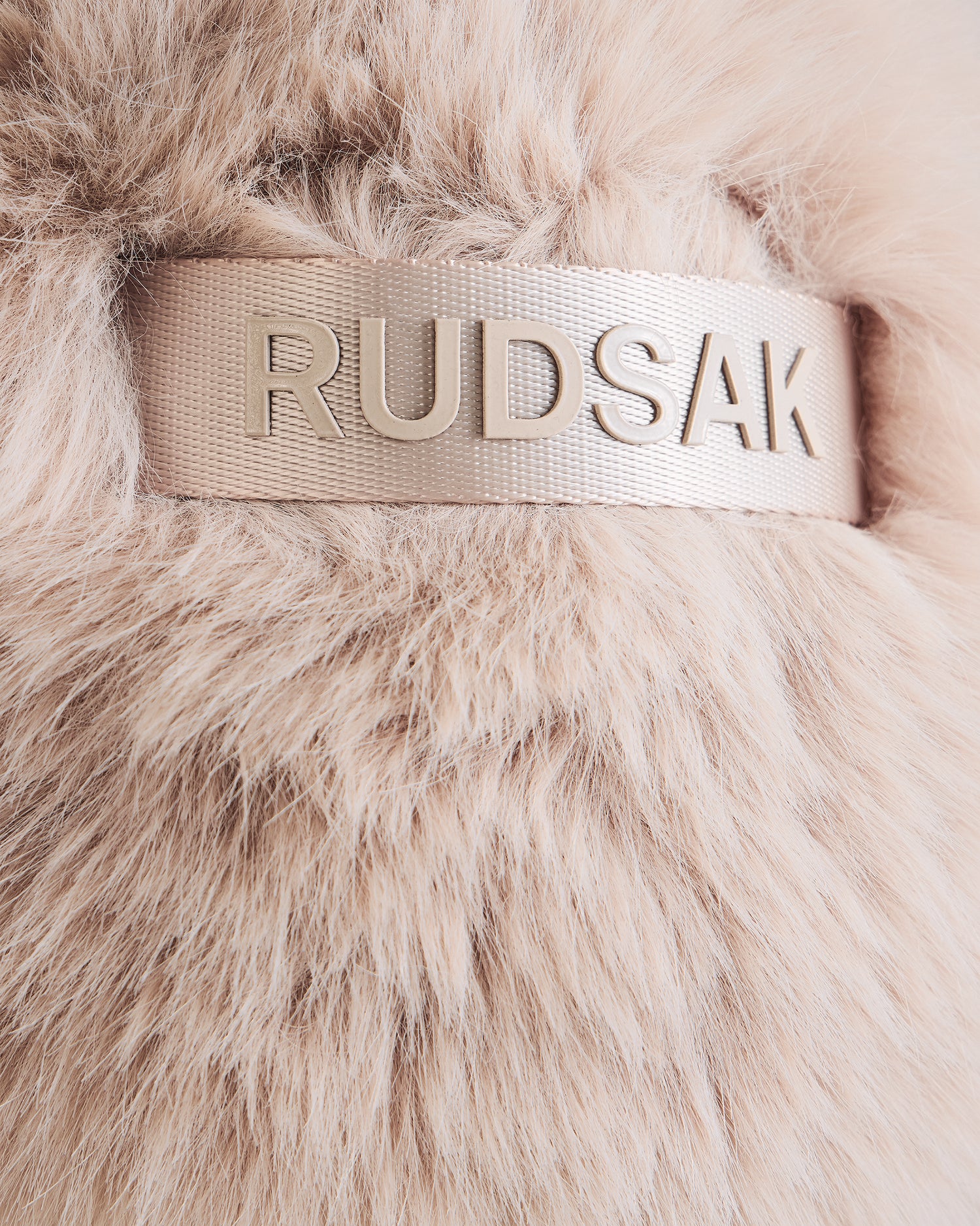 Close-up of soft, light beige faux fur from Rudsak’s DEMI - L425539 Vanilla women’s aviator hat, featuring a shiny beige band with raised metallic RUDSAK lettering—an elegant detail on this chic winter accessory.