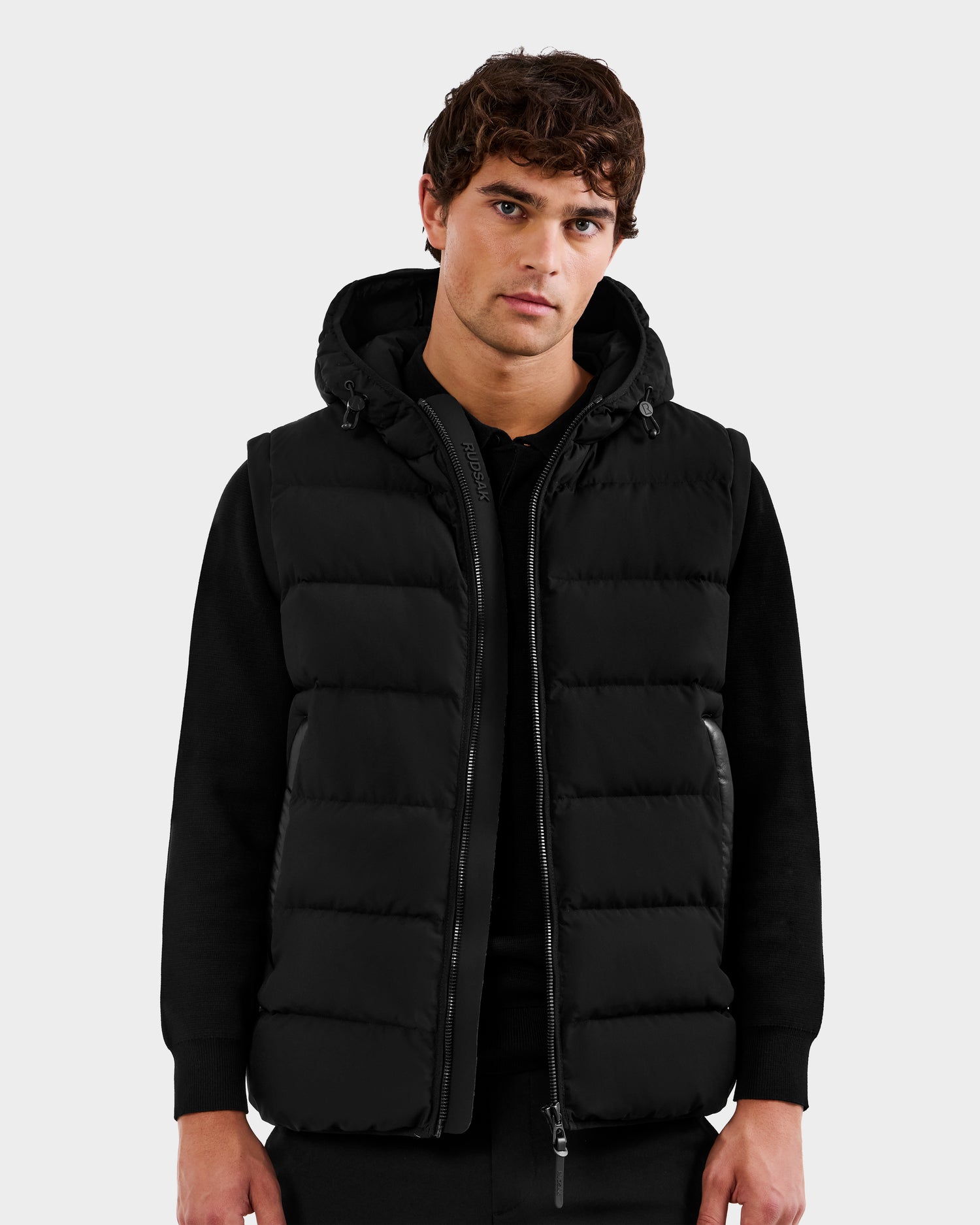 A young man with short, wavy brown hair wears the Rudsak DEMIAN M-R - M123504 Black men’s down puffer vest, made from sustainable recycled fabric, over a black long-sleeve shirt, standing against a light gray background.