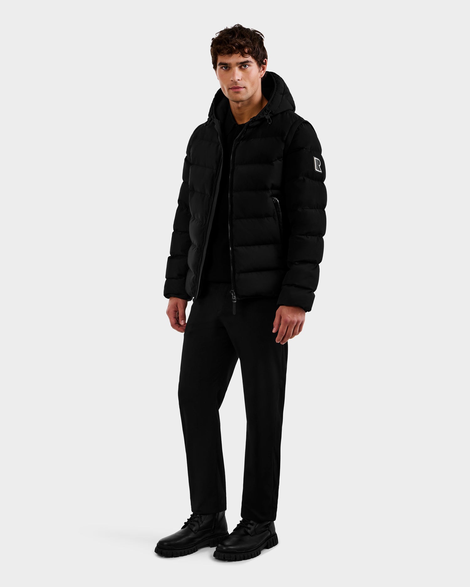 A man faces forward, wearing the Rudsak DEMIAN M-R - M123504 Black men's down puffer made from sustainable recycled fabric, paired with black pants and shoes. His hands rest at his sides against a plain light gray background.