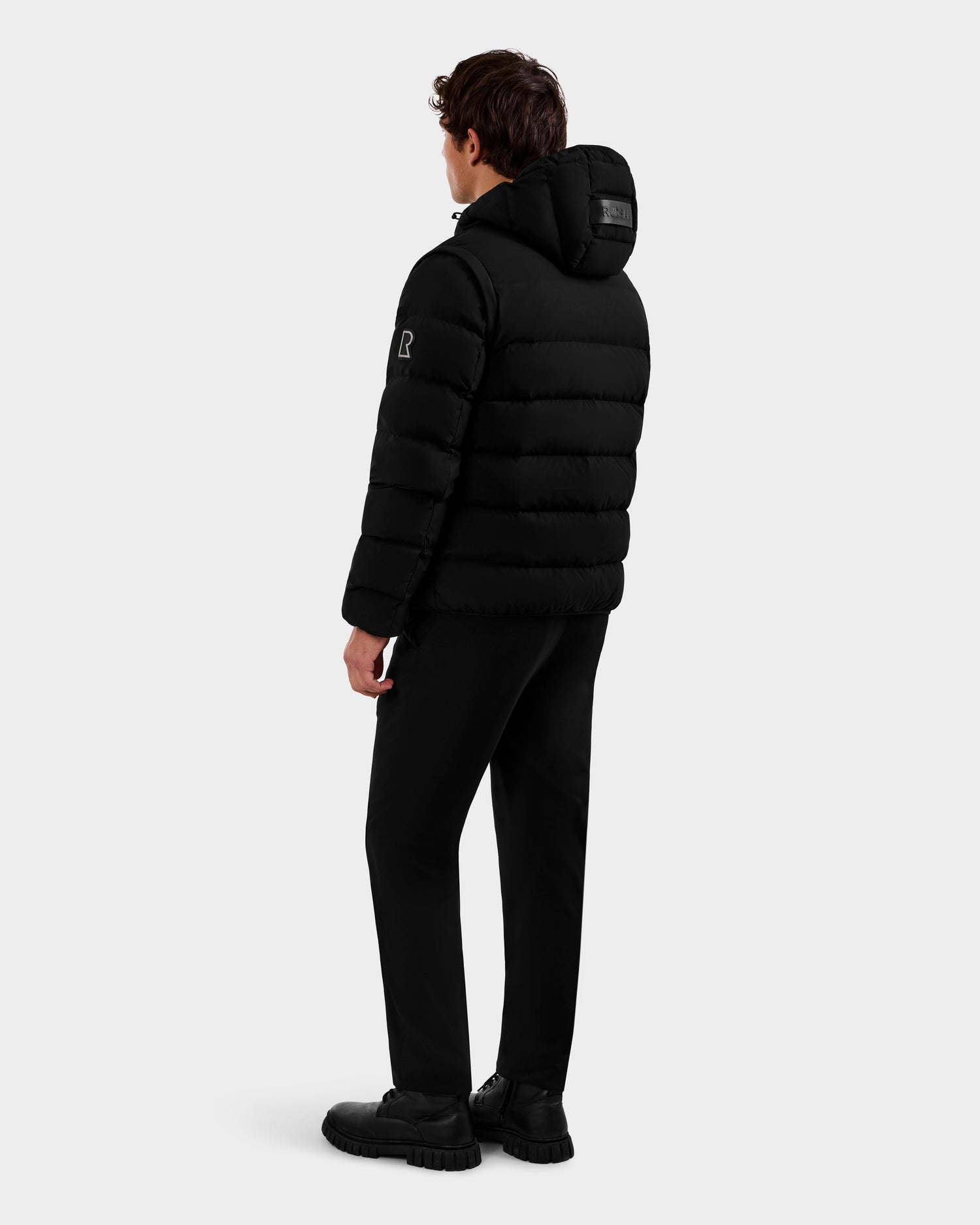 A person faces away, wearing the Rudsak DEMIAN M-R - M123504 Black men's down puffer jacket with a hood, paired with black pants and black shoes, set against a plain light gray background.