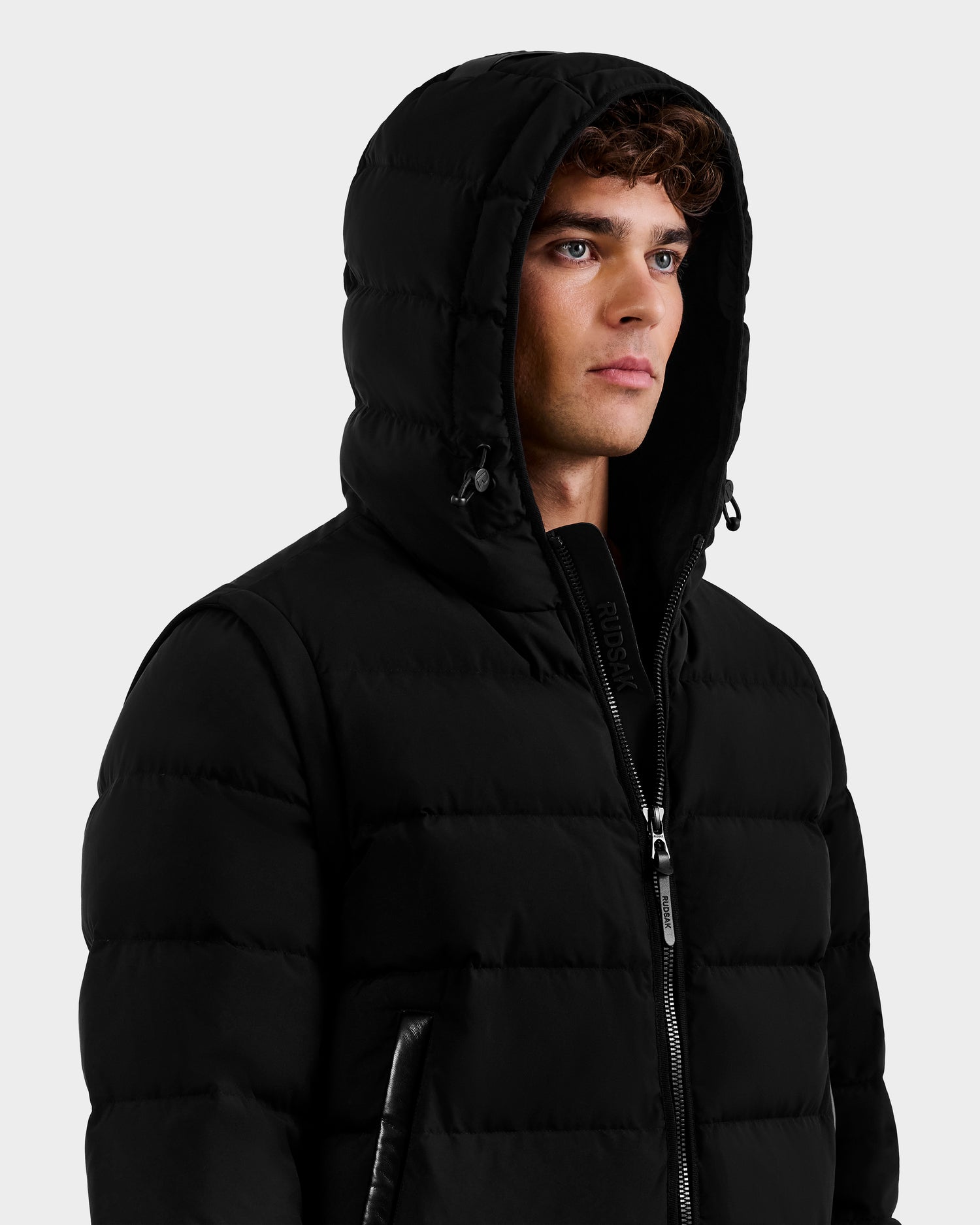 A person wearing the Rudsak DEMIAN M-R - M123504 Black men's down puffer jacket with a hood and front zipper, gazing sideways against a plain light background. Made from sustainable recycled fabric for eco-friendly style.