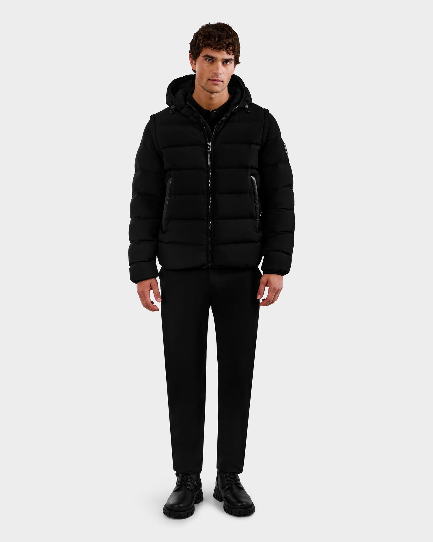 A man stands facing forward, wearing the Rudsak DEMIAN M-R - M123504 Black men's down puffer jacket, paired with black pants and shoes, set against a plain light gray background.