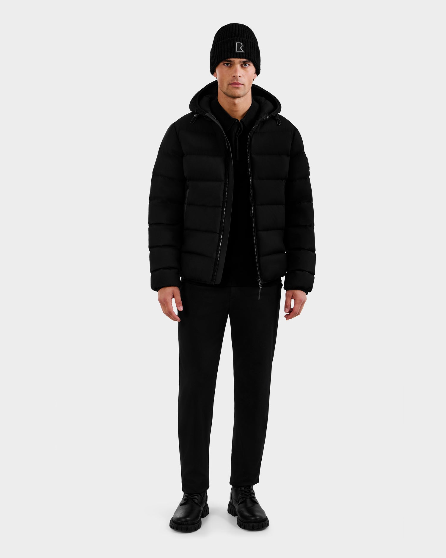 A man faces forward wearing the Rudsak DEMIAN M - M123507 Black men’s down puffer jacket, along with a black beanie, shirt, pants, and shoes made from sustainable recycled fabric, set against a plain white background.
