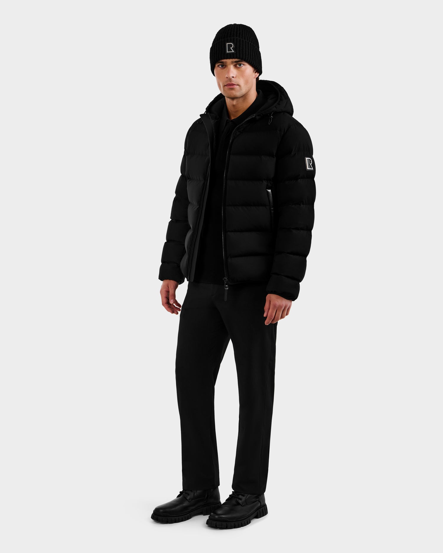 A person wearing the Rudsak DEMIAN M - M123507 Black men’s down puffer stands against a plain white background, styled with a black beanie, black pants, and black shoes.