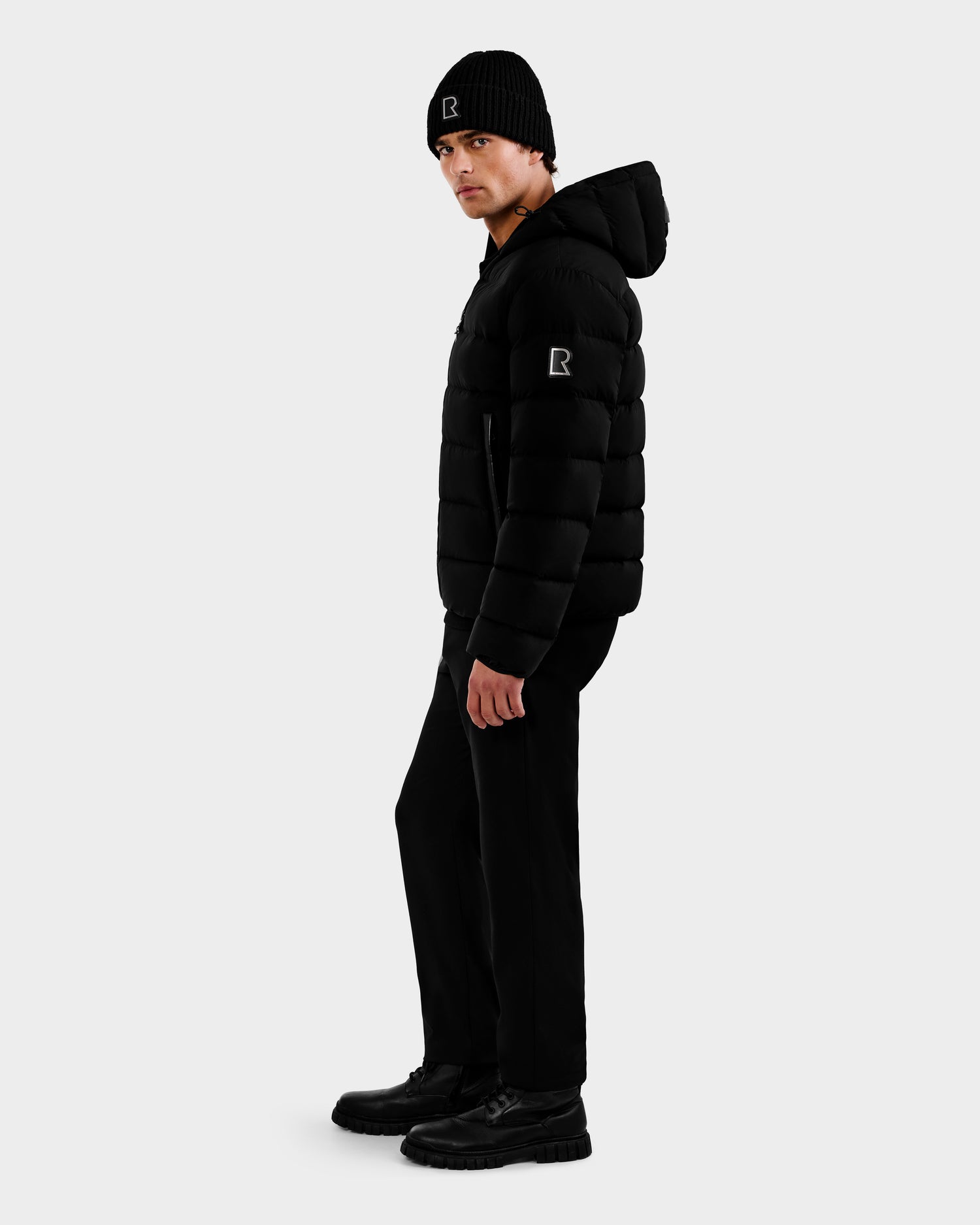 A man stands sideways in a Rudsak DEMIAN M - M123507 Black hooded down puffer made from sustainable recycled fabric, paired with a black beanie, pants, and shoes against a plain light gray background.