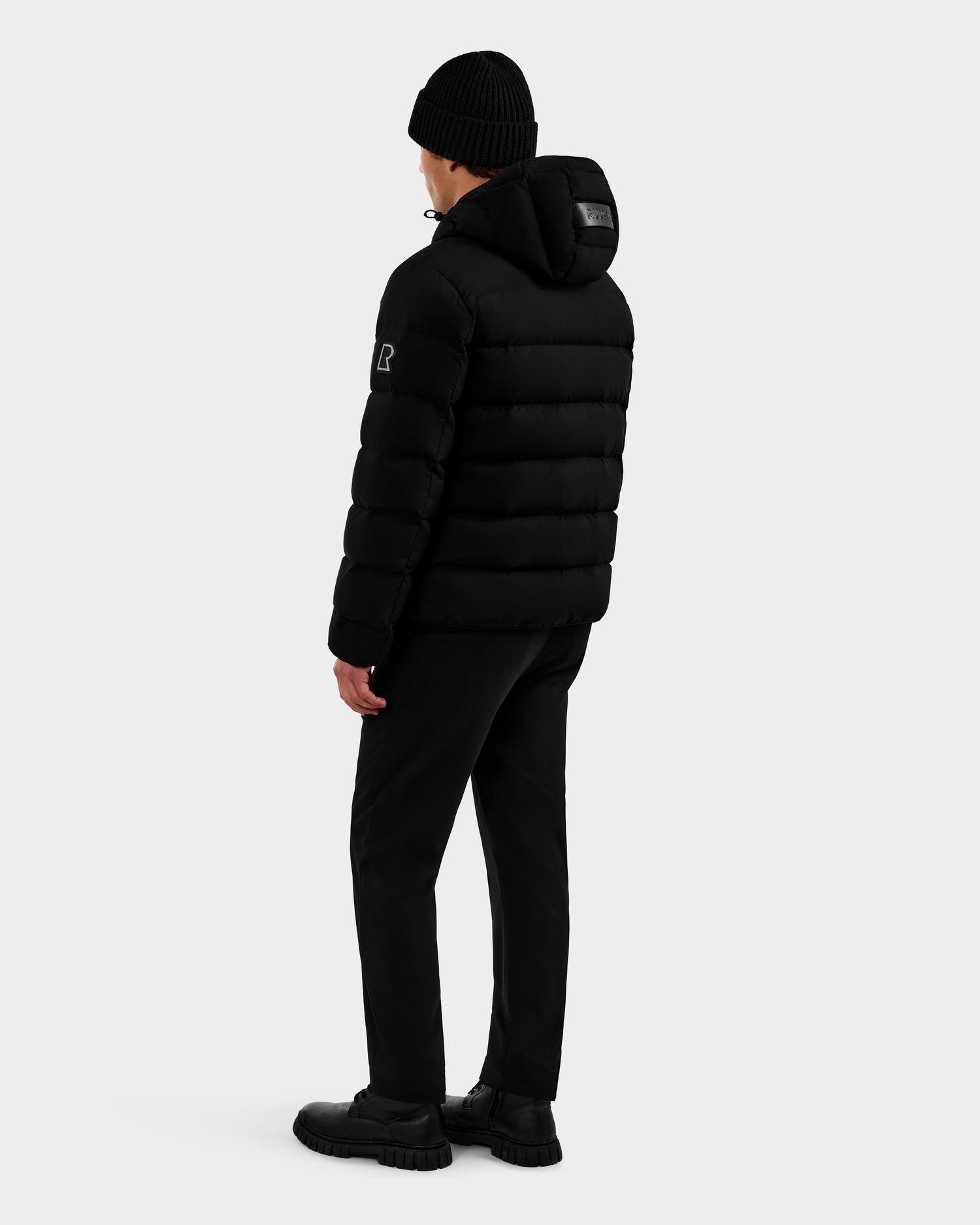 A person wears the Rudsak DEMIAN M - M123507 Black men’s down puffer with sustainable recycled fabric, black beanie, pants, and shoes, standing turned to the side against a plain light gray background.