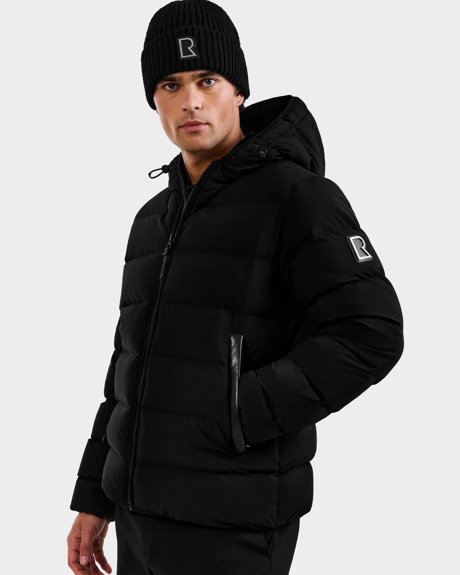 A man wears the Rudsak DEMIAN M - M123507 Black beanie and hooded down puffer jacket with zip front and R patch, made from sustainable recycled fabric, standing against a plain light background.