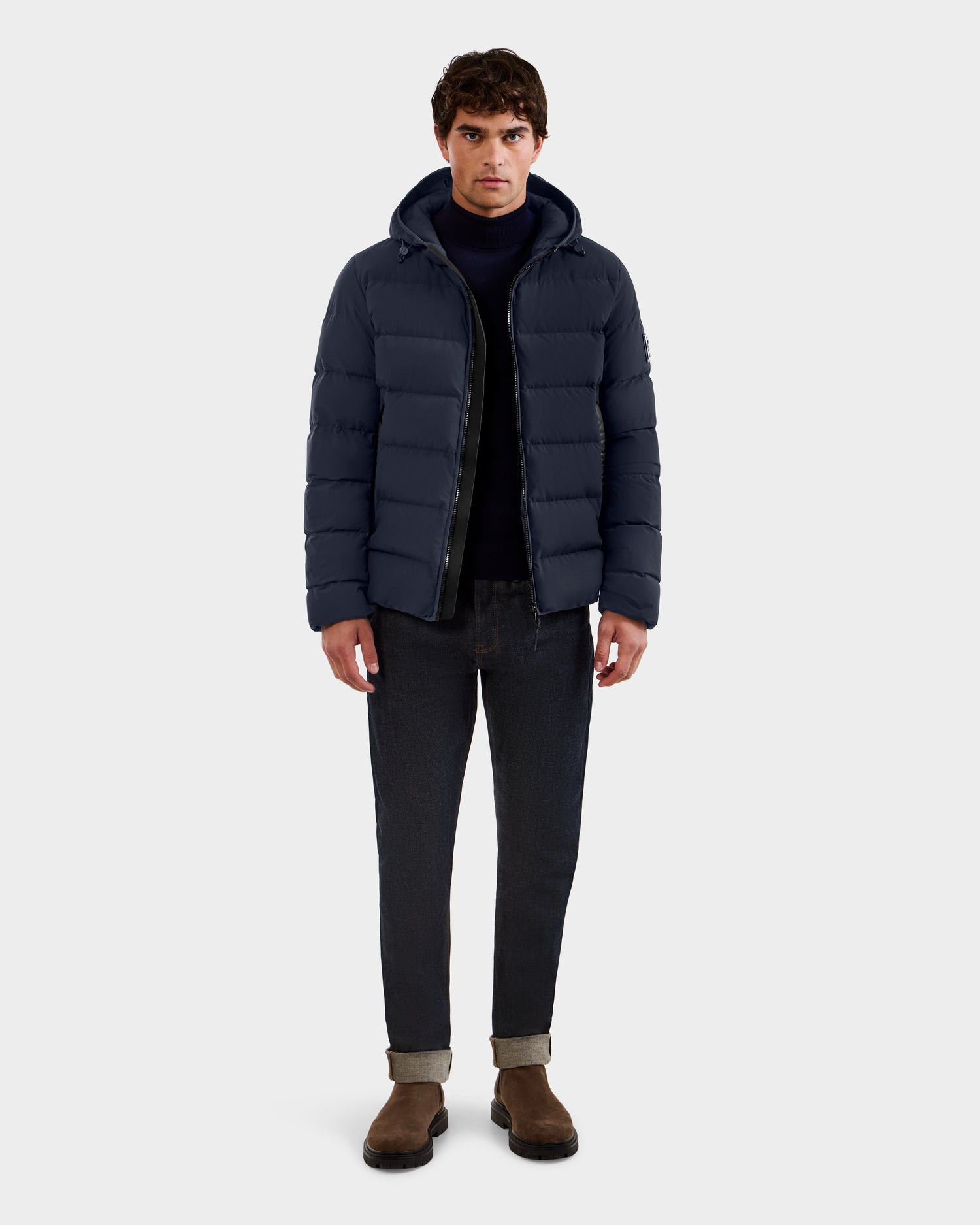 A man stands facing forward against a plain light gray background, wearing the Rudsak DEMIAN M - M123507 Navy men’s down puffer jacket with sustainable recycled fabric, paired with dark jeans, brown boots, and a dark sweater.