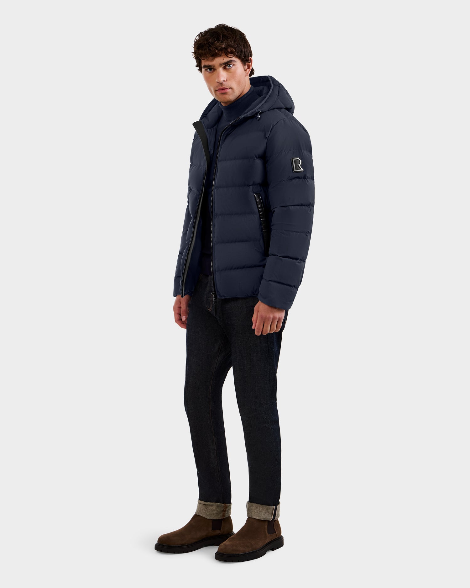 A man wearing the Rudsak DEMIAN M - M123507 Navy down puffer jacket made from sustainable recycled fabric, paired with dark cuffed jeans, a navy hoodie, and brown Chelsea boots stands against a plain background with a neutral expression.