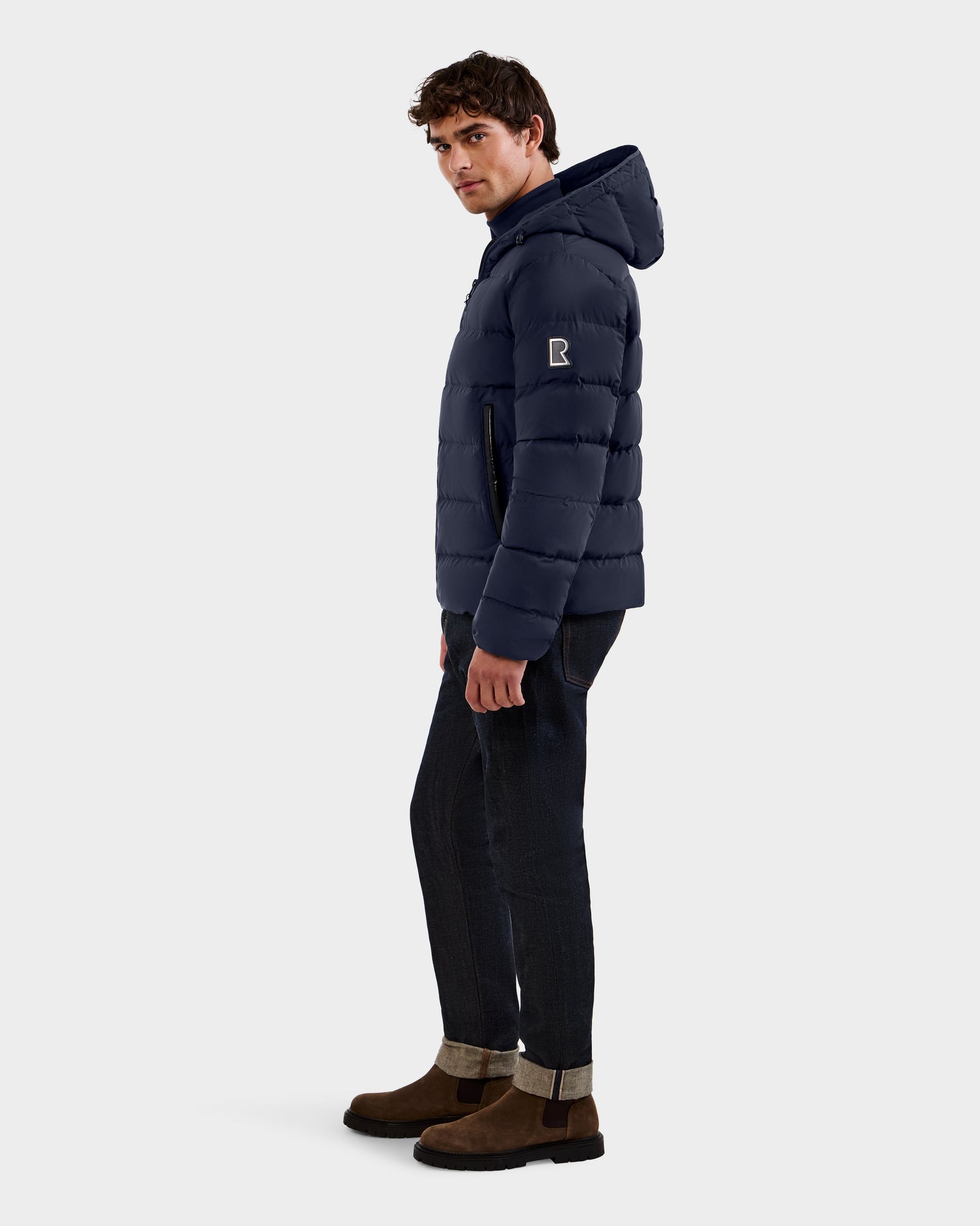 A man stands sideways in the Rudsak DEMIAN M - M123507 navy men’s down puffer jacket with a hood, made from sustainable recycled fabric, paired with dark cuffed jeans and brown suede boots against a plain white background.